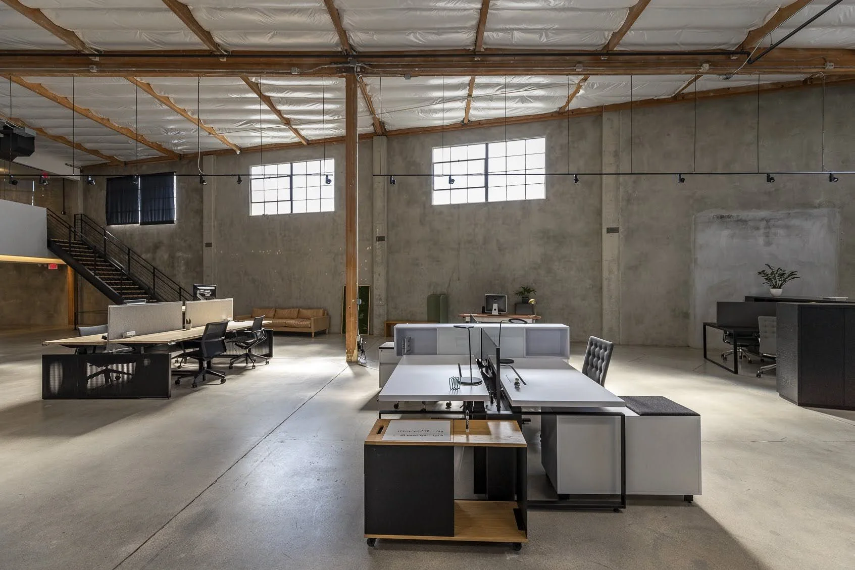 Open plan office with modern furniture, concrete walls, and large high windows, featuring desks, chairs, plants, and a staircase.