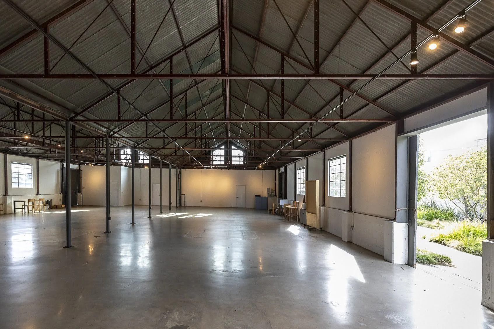 Large empty industrial-style space with high metal ceiling and big windows, sunlight streaming in, and some chairs and furniture along the walls.