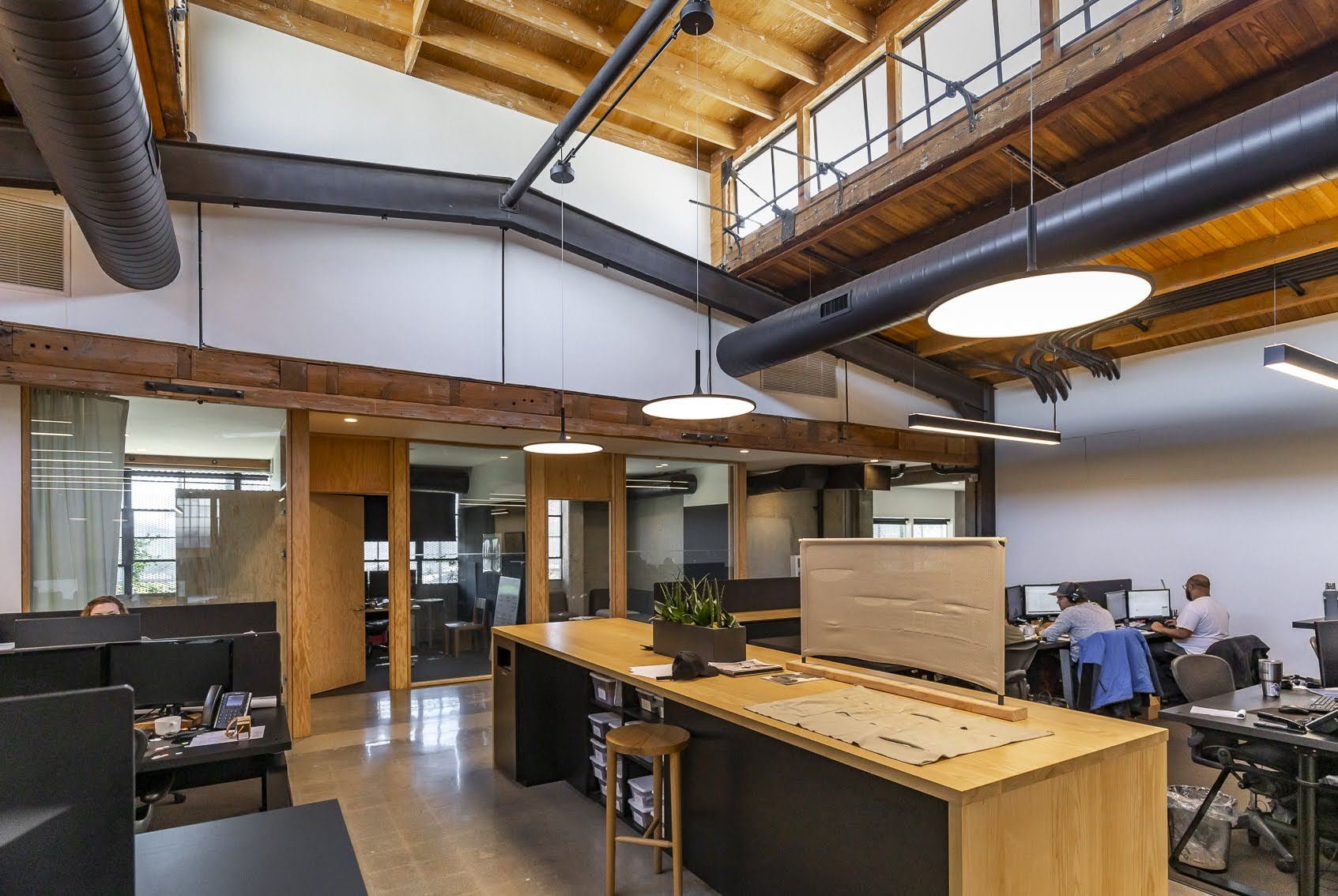 Office space with exposed wooden beams, modern lighting fixtures, and employees working at desks.