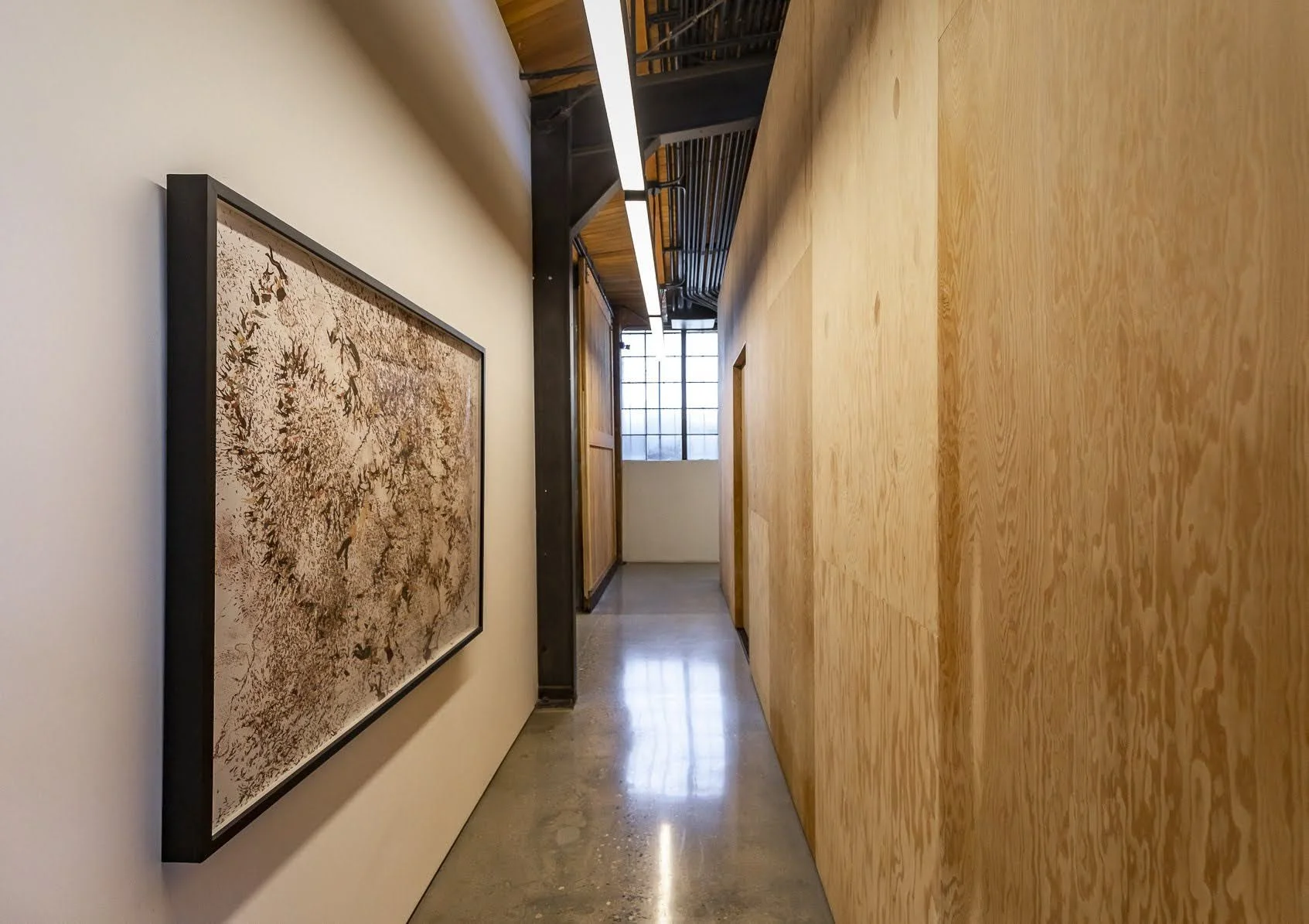 A hallway with a minimalist design, white and wooden paneled walls, polished concrete flooring, and a large window at the end allowing natural light. There is a framed artwork with an abstract pattern on the left wall and architectural elements like 