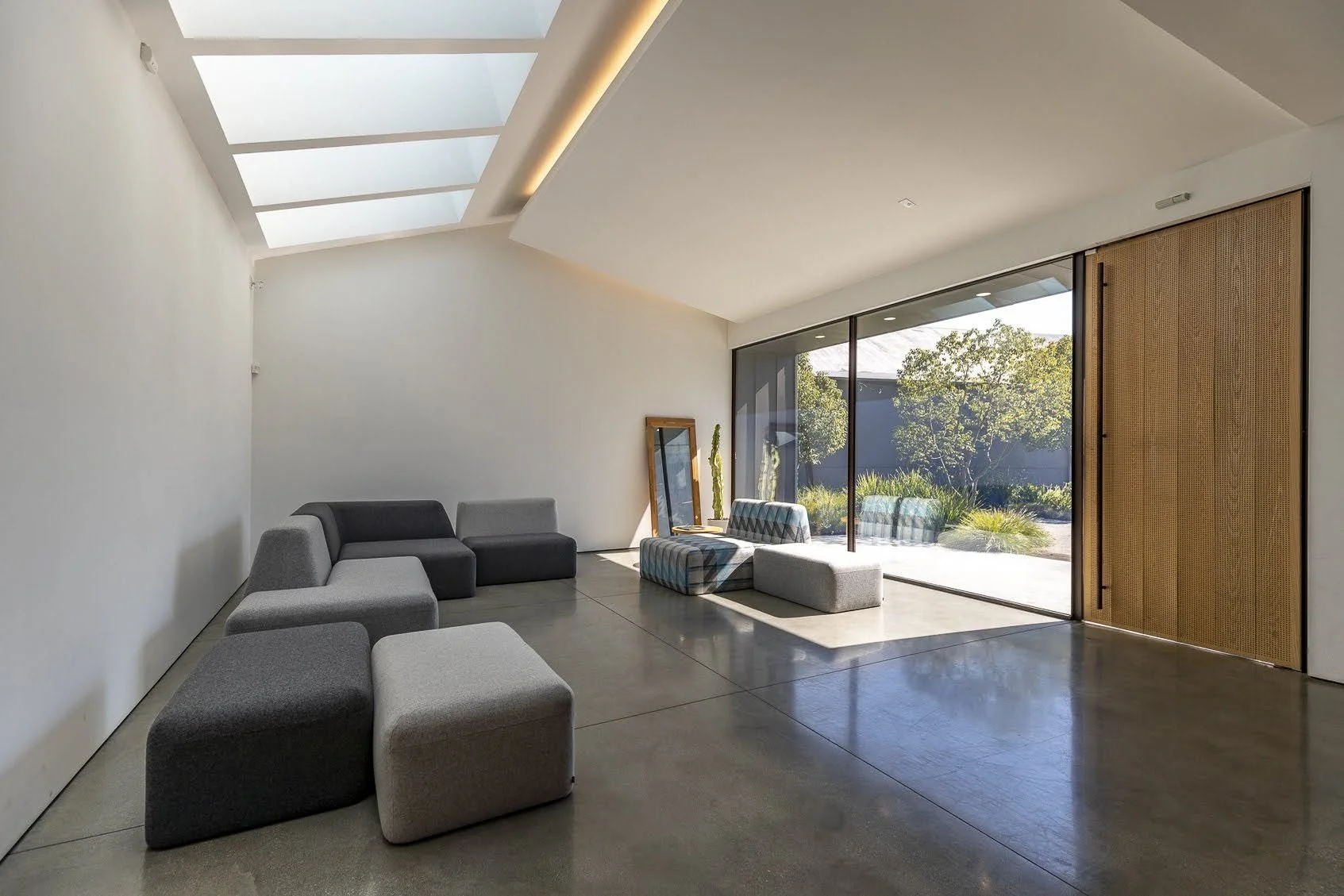 Modern living room with minimalist furniture, large glass sliding doors revealing outdoor garden, and a skylight letting in natural light.
