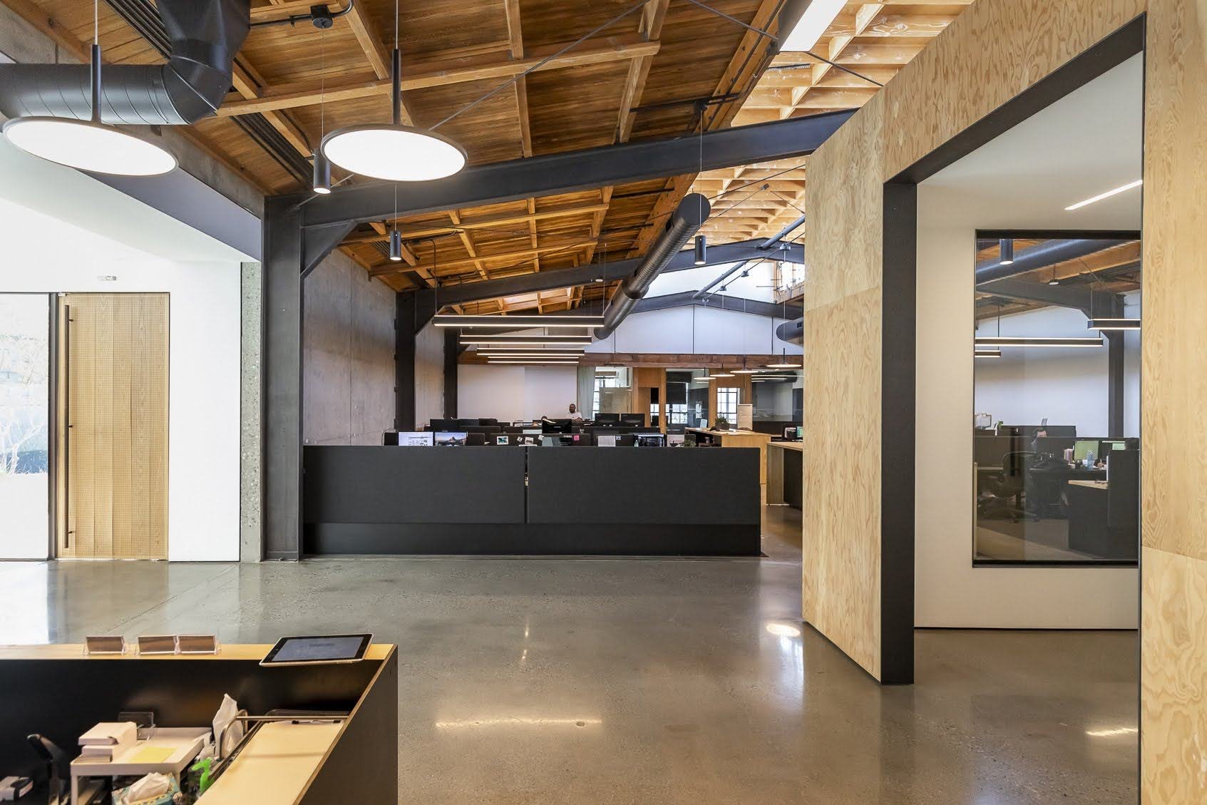 Modern office reception area with wood and black accents, open ceiling with exposed wood beams and black ventilation ducts, front desk with computer monitors, and workspaces with desks and chairs in background.