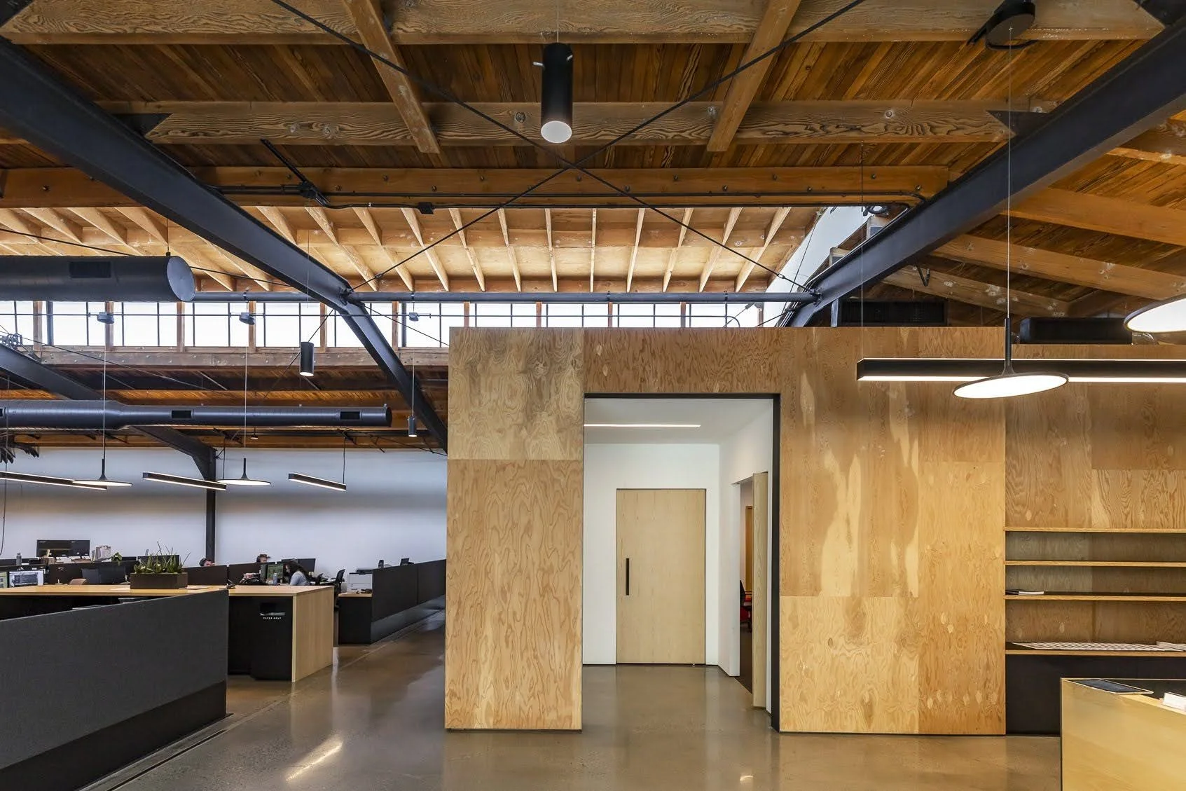 Modern office interior with wood panel walls and ceiling, exposed black structural beams, and suspended linear lighting.