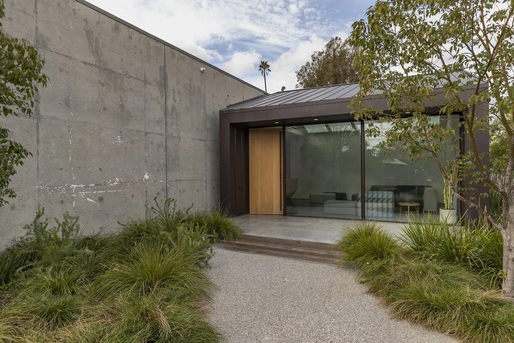 Modern house with large glass door, concrete exterior wall, and landscaped front yard with plants and a tree