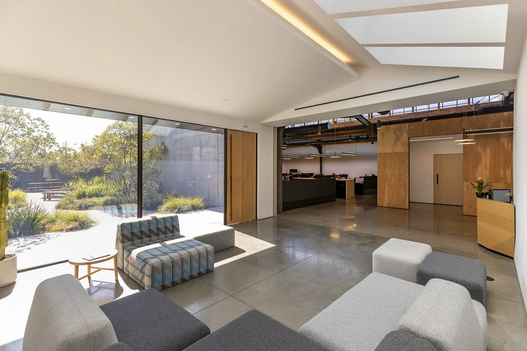 Modern office lobby with large glass windows, gray and plaid couches, potted plants, and natural lighting.