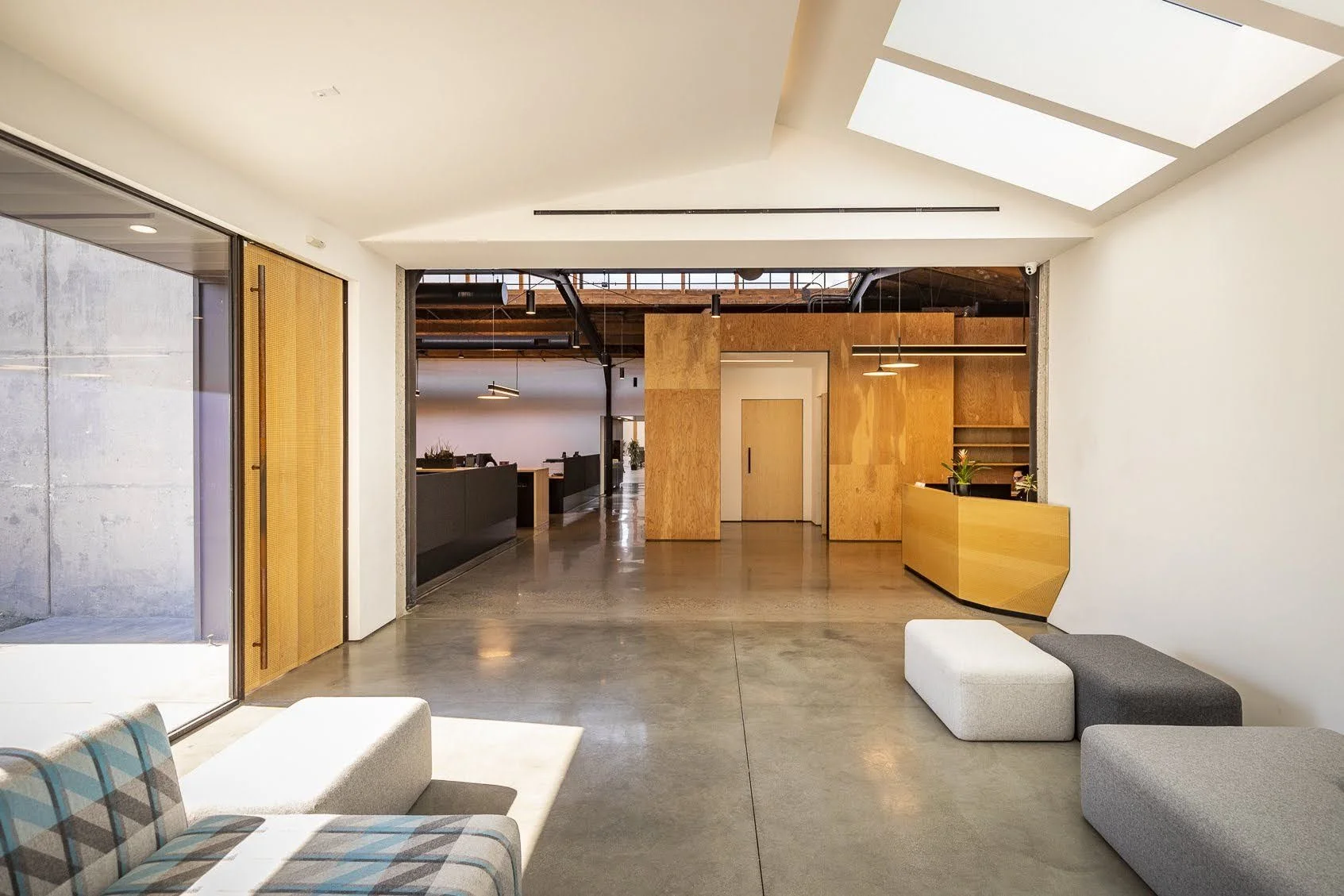 Modern office lobby with minimalist furniture, wood accents, and a polished concrete floor, illuminated by natural light from skylights.