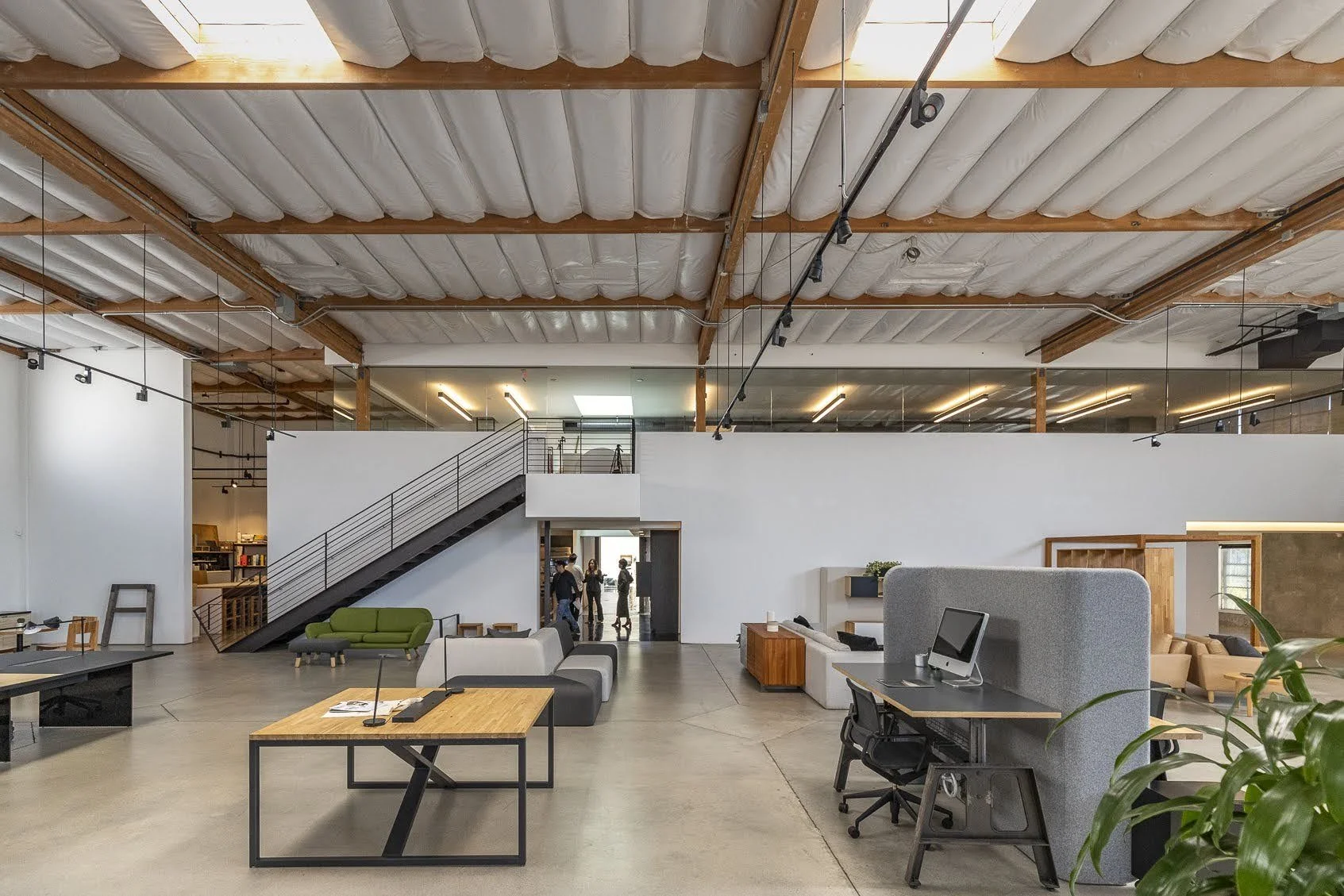 Modern open-concept office space with sitting areas, desks with computers, and a staircase leading to an upper level. People are visible near the staircase and in the background. The ceiling features exposed beams and insulation, with track lighting.