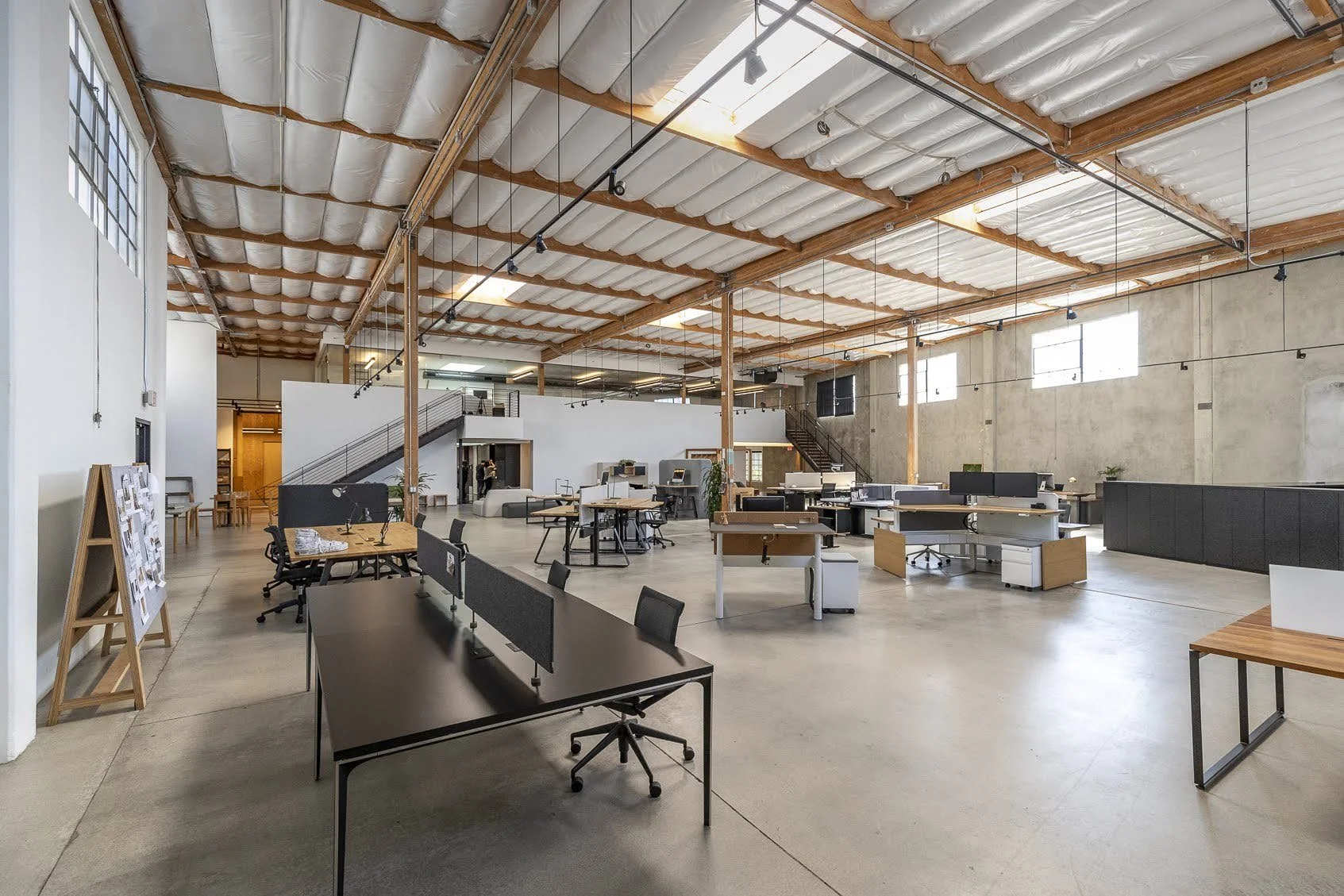 Spacious modern office with high ceilings, exposed wooden beams, and large windows allowing natural light. The open layout includes desks, chairs, and workstations.