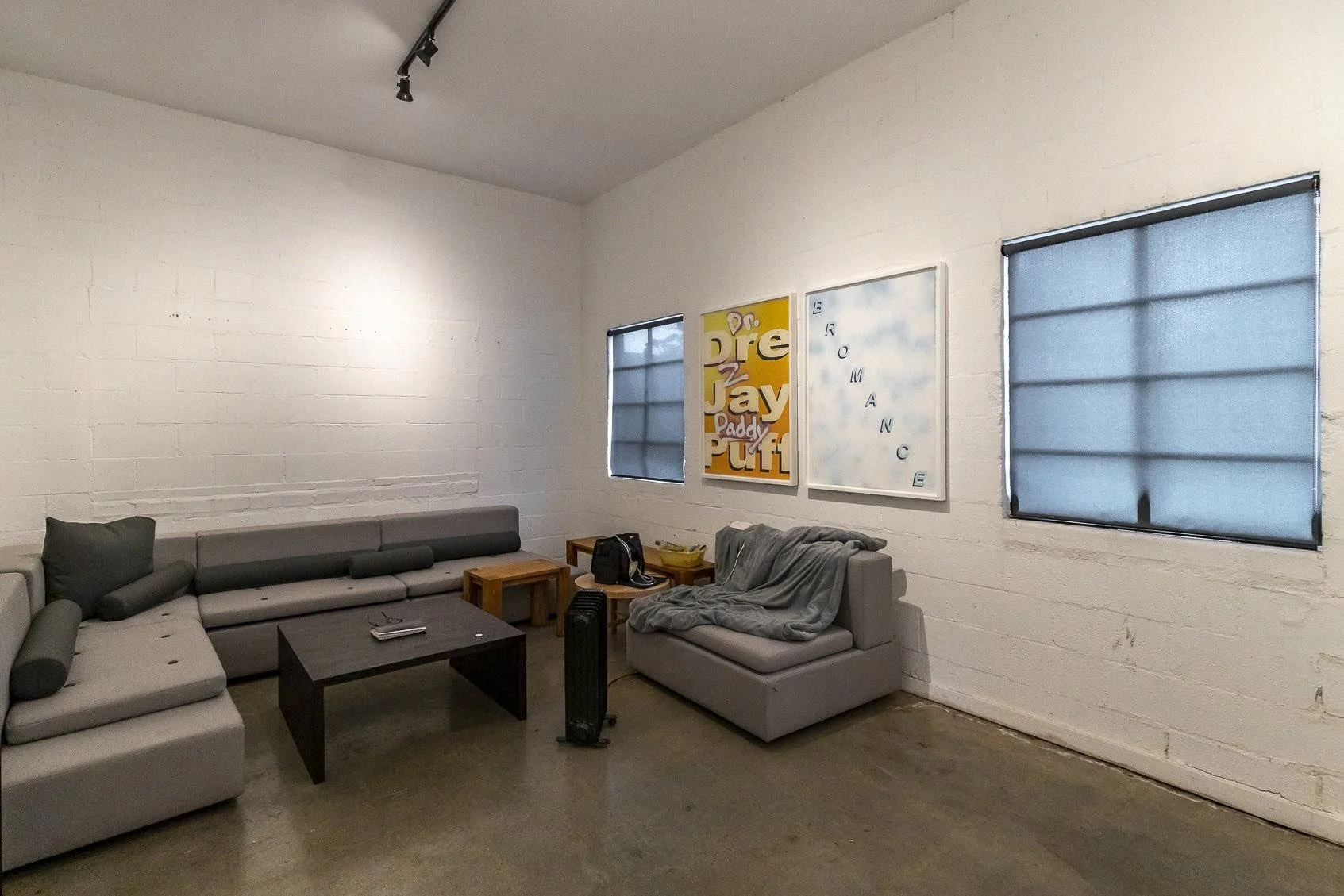 A living room with white brick walls and three windows with blue blinds. There is a gray sectional sofa with a pillow and a blanket, a small wooden side table, a black portable fan, and three framed wall art pieces with colorful designs and text.