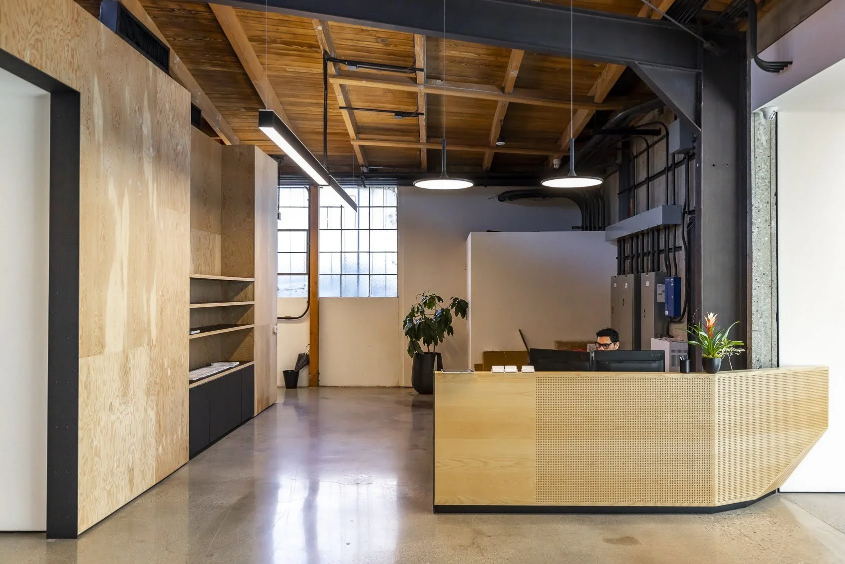 Modern office reception area with a wooden desk, a person working at a computer, plants, large frosted windows, and exposed wooden ceiling beams.