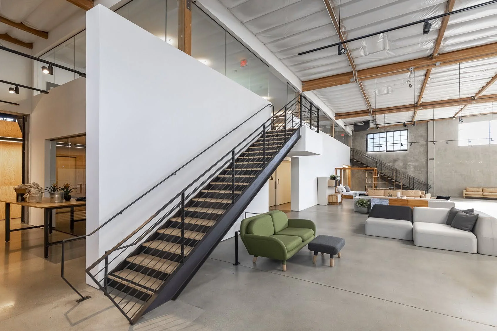 Modern office lobby with two staircases, glass partitions, and contemporary furniture including green, gray, and beige sofas.