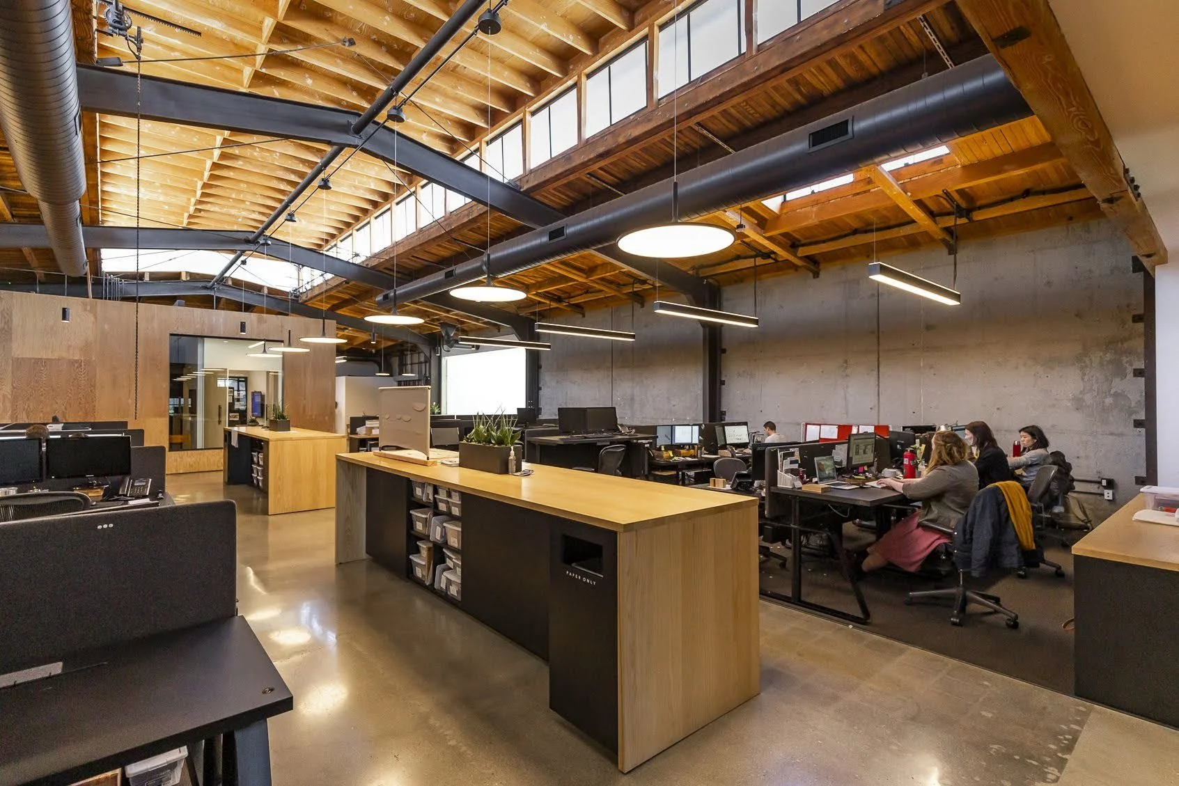 An open-plan office with high wooden ceilings, large windows, and multiple employees working at desks.
