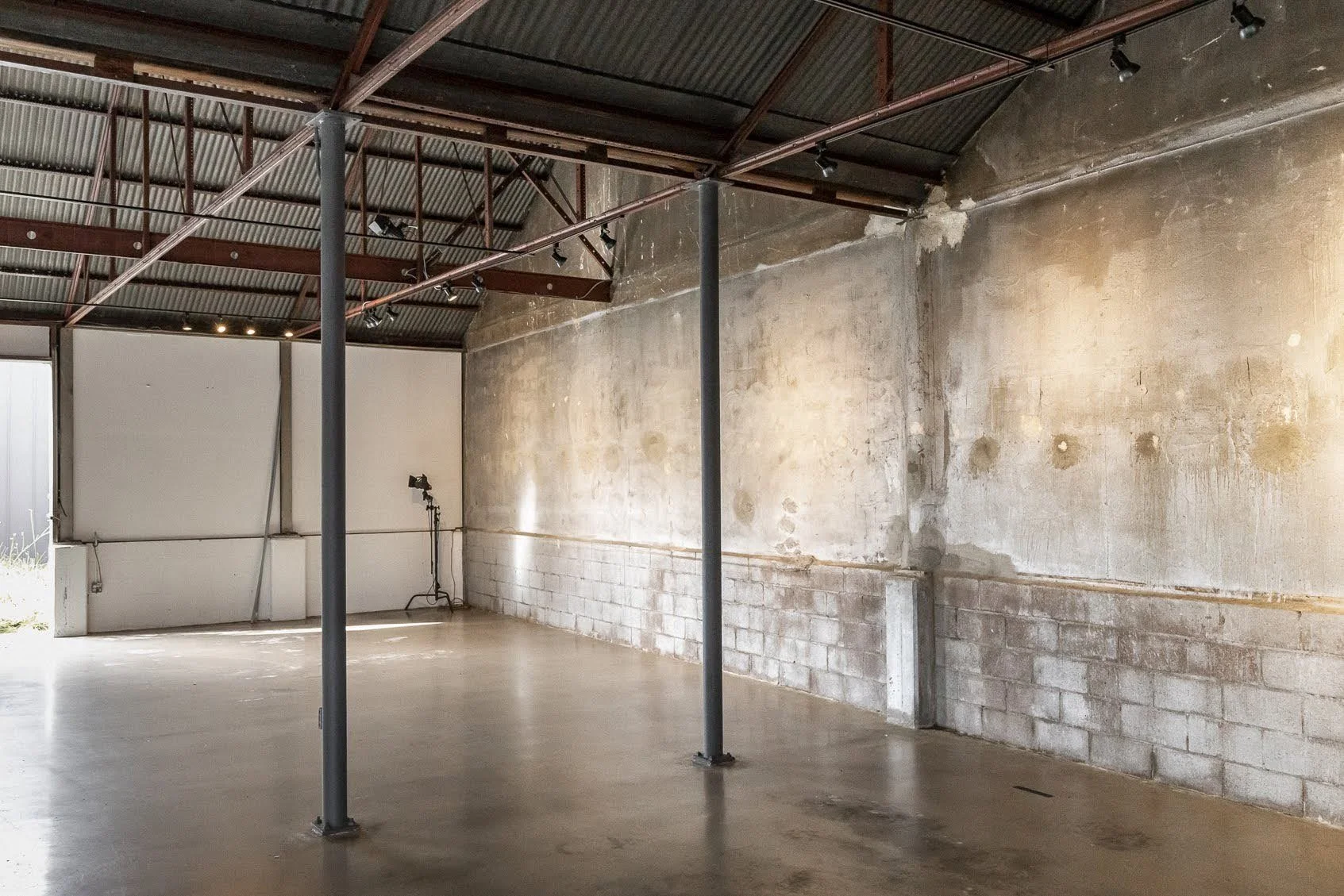 Empty industrial-style interior space with concrete flooring, unfinished walls, and exposed ceiling beams with lighting.