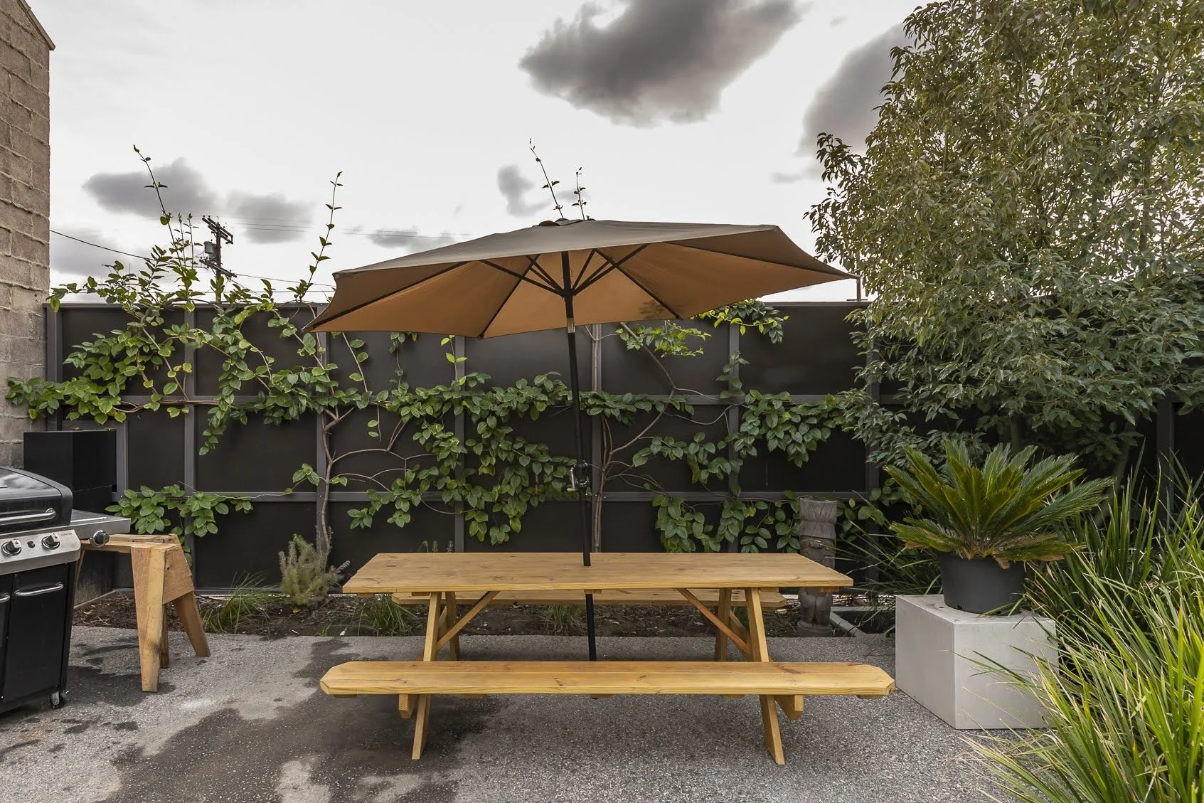 Outdoor patio with wooden picnic table, large tan umbrella, black privacy fence, potted plants, and cloudy sky.
