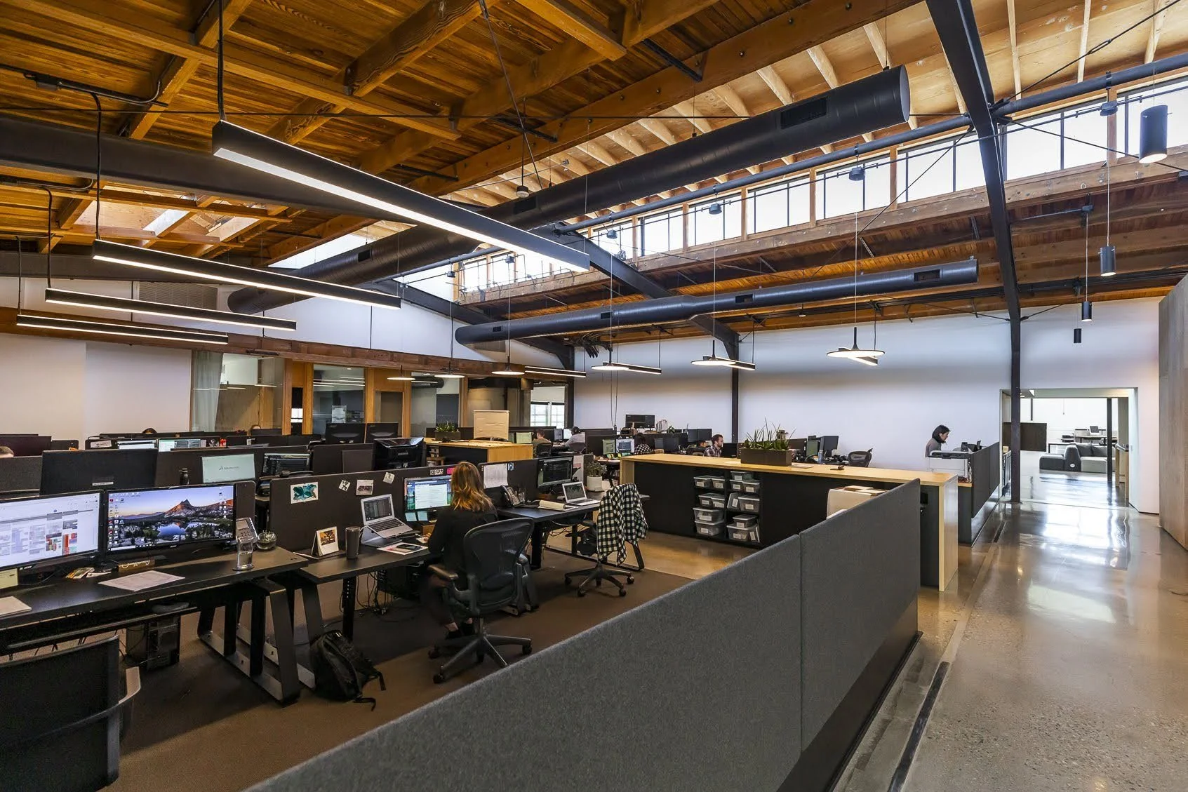 Open-plan modern office with workers at desks, exposed wooden ceiling, and large windows letting in natural light.