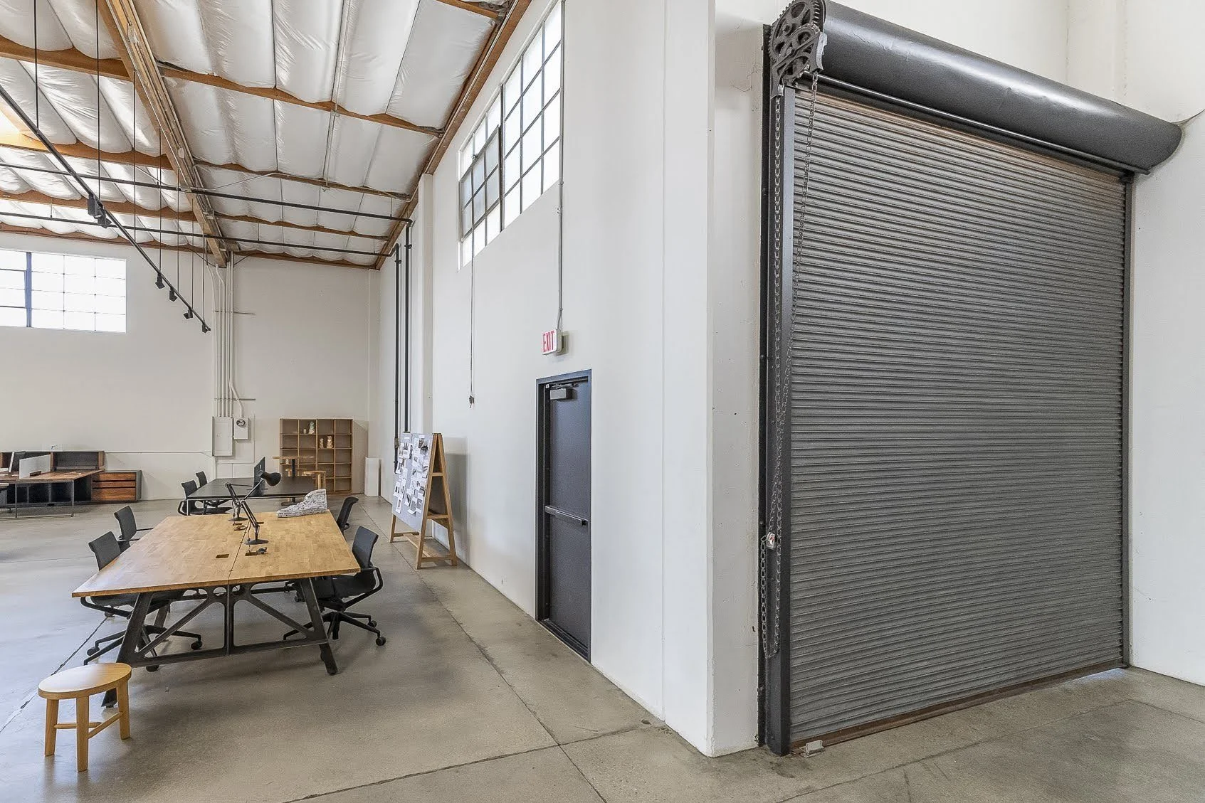 Interior of a warehouse or industrial space with high ceilings, a large rolling metal door, a table and chairs, window panels, and a small stool.