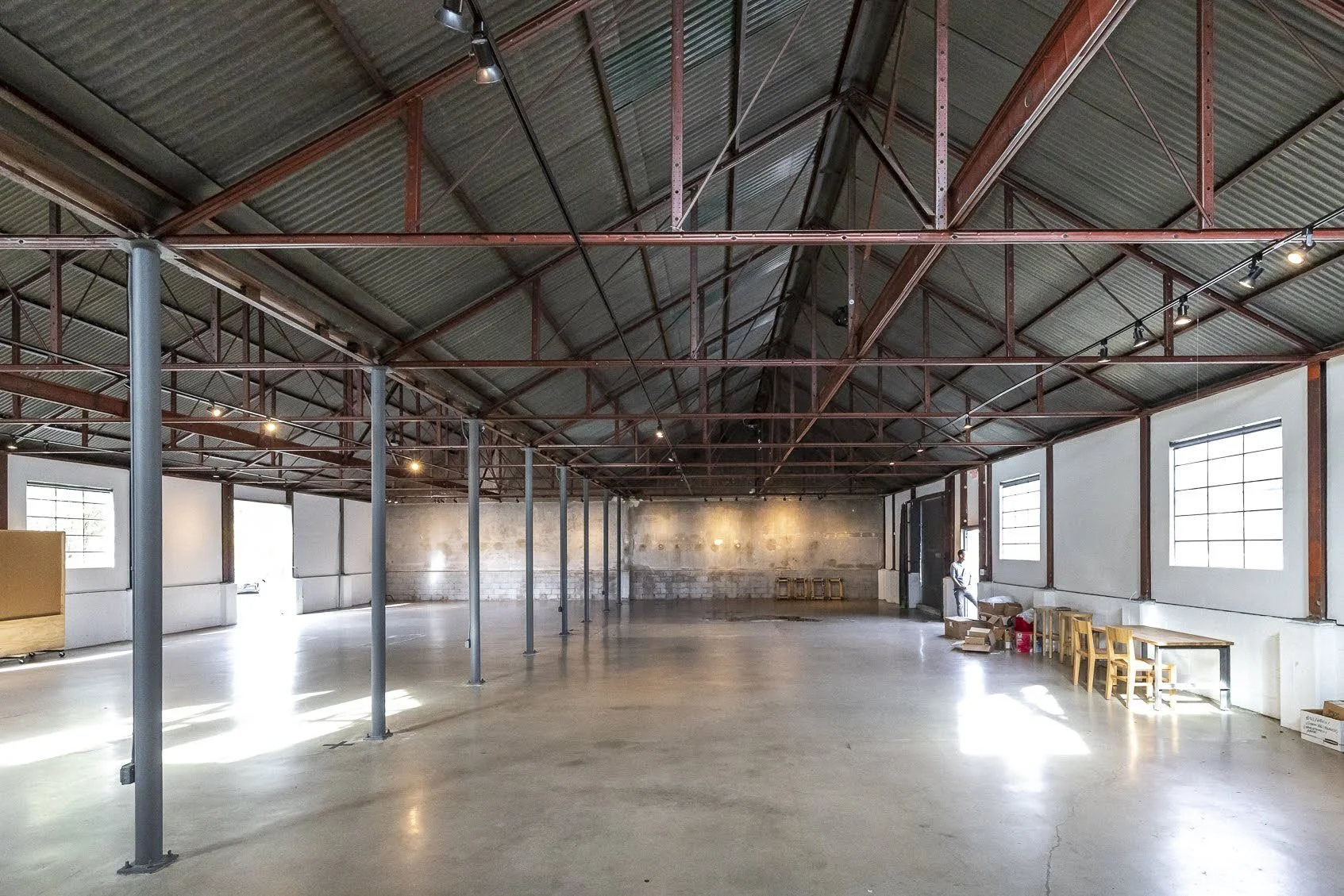 An empty industrial-style interior space with high ceiling, exposed brick and metal beams, concrete floor, and large windows letting in natural light.