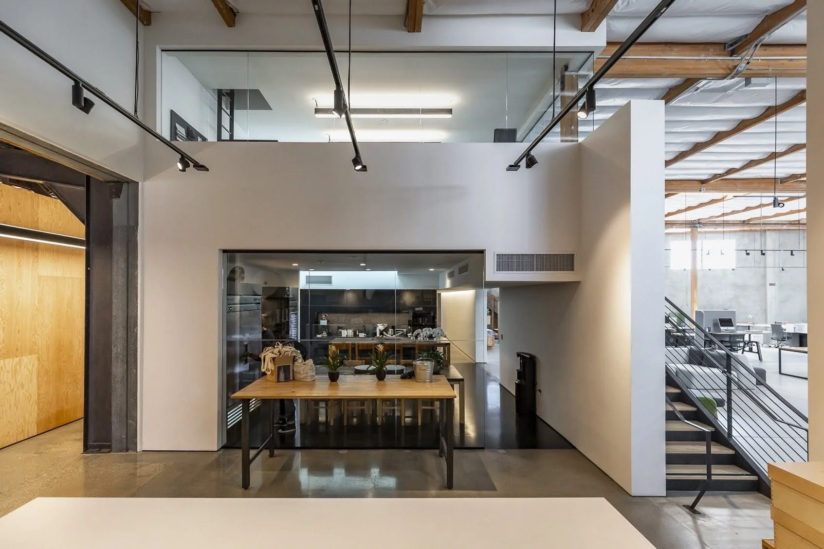 Modern interior of a cafe or workspace with a high ceiling, exposed wooden beams, track lighting, and a glass-enclosed kitchen area with a wooden table and potted plants in front.