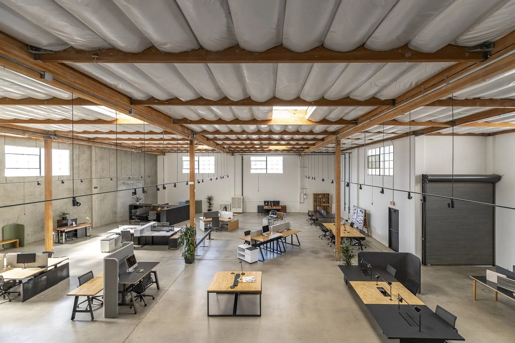 Open-plan office space with desks, chairs, and computers, featuring exposed ceiling beams and windows.