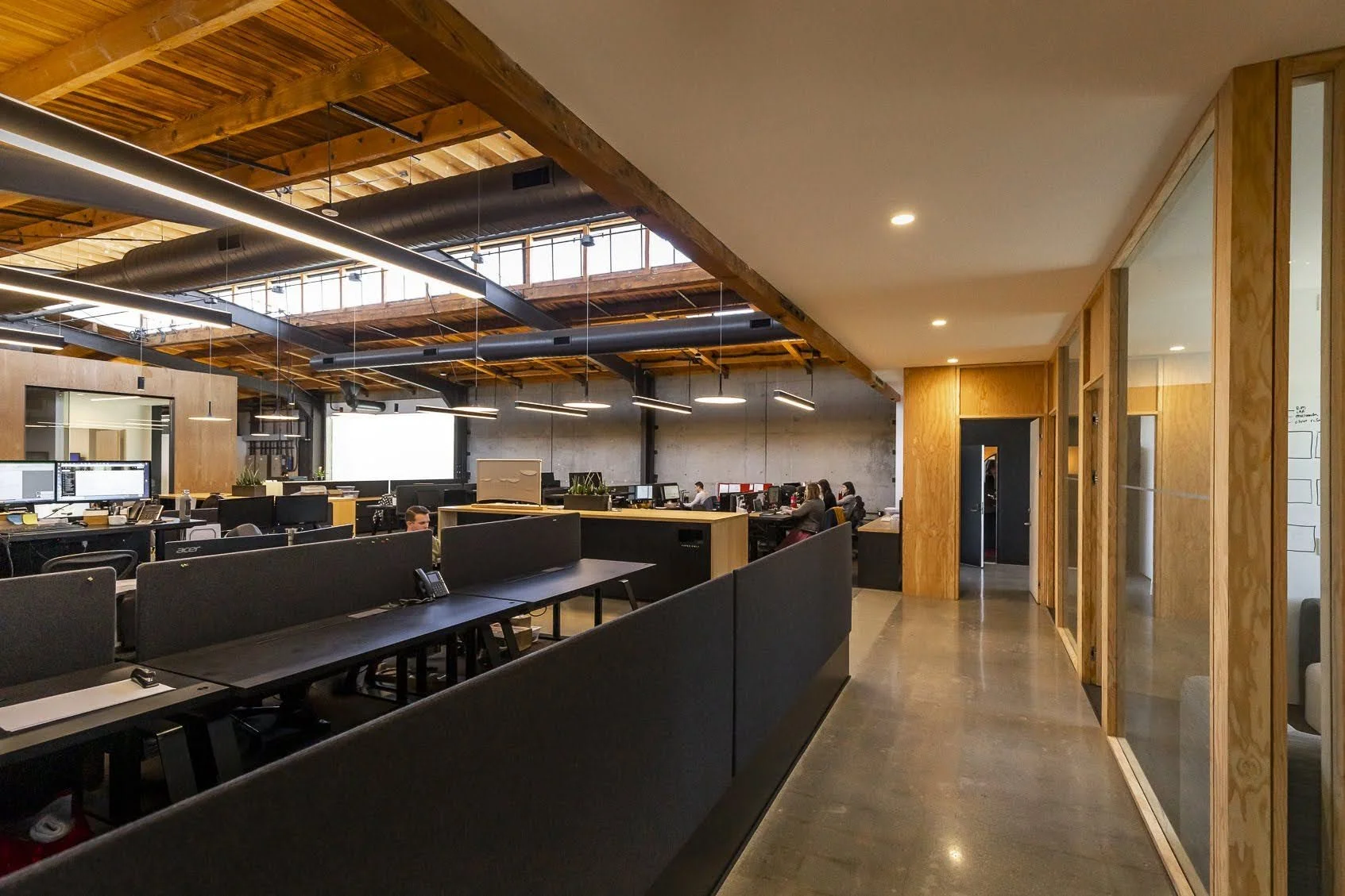Modern open-plan office with black desks, computer monitors, and staff working, featuring wooden beams, ceiling windows, and glass partition walls.