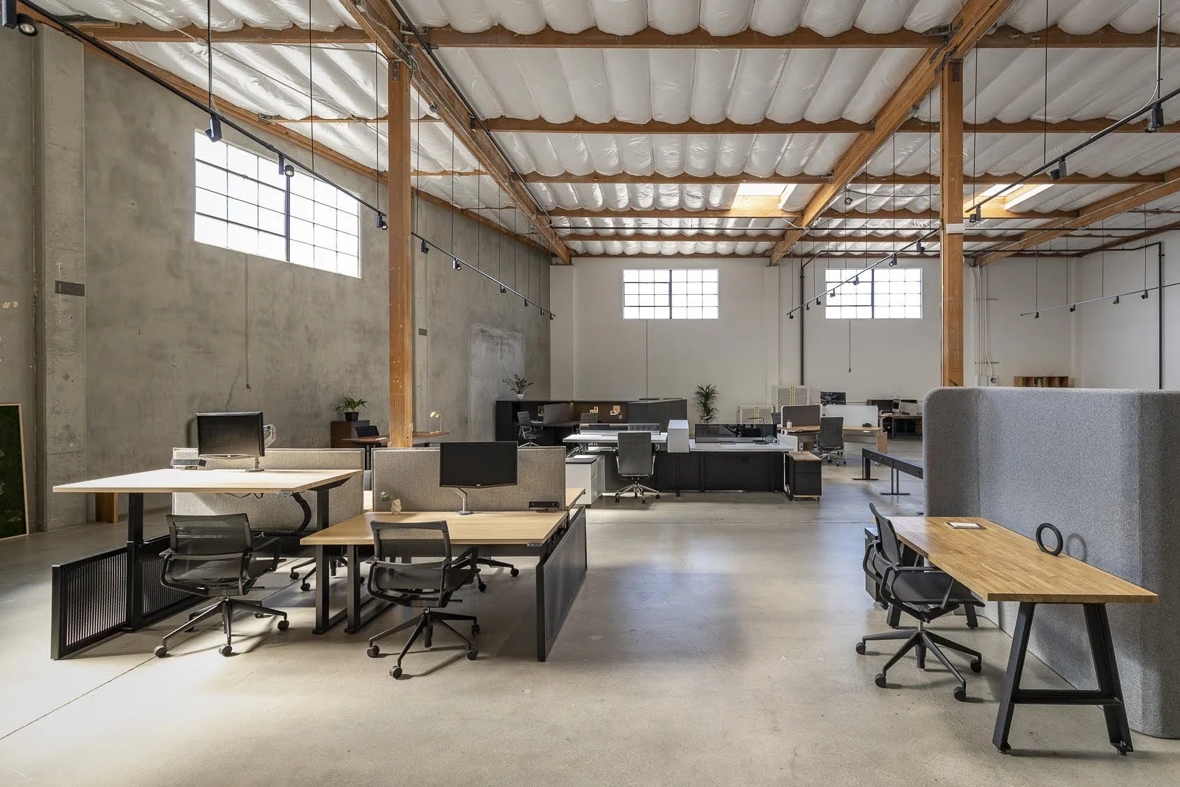 Open office space with modern desks, office chairs, computers, and plants, featuring high ceilings with wooden beams and large windows.