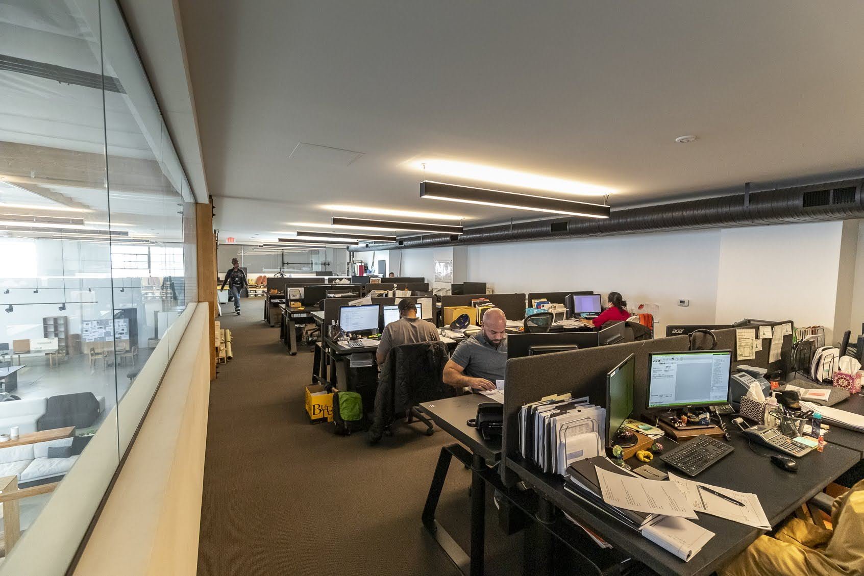 Office workspace with multiple desks, computers, and employees working, viewed from a hallway with a glass wall.