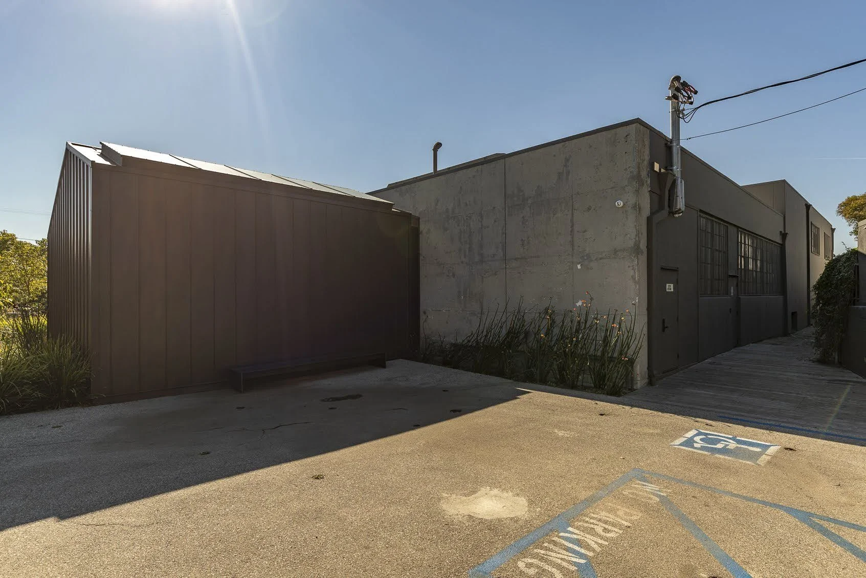 A modern building with a mix of industrial and minimalist design, featuring concrete and metal walls, a small ramp, and a designated handicapped parking space in the foreground, under clear blue skies.