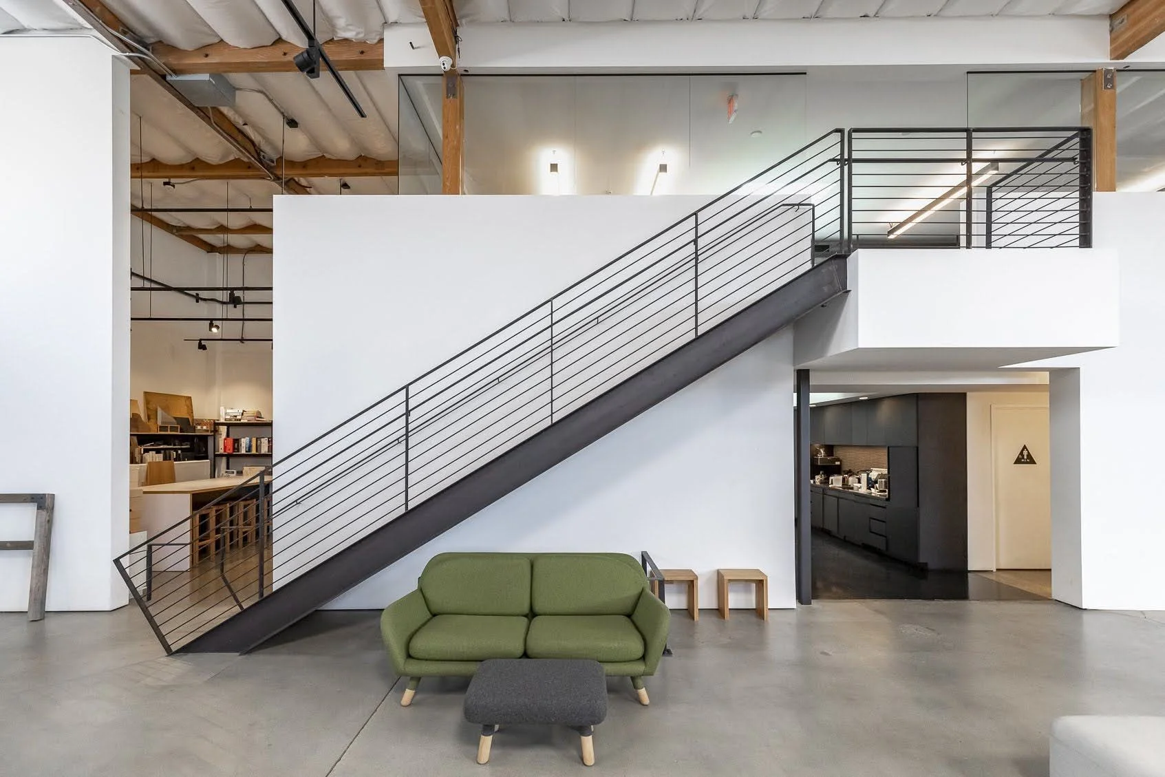 Modern interior workspace with a green sofa, black ottoman, and a staircase with black railings leading to an upper level, exposed wooden ceiling beams, and a visible kitchen area.