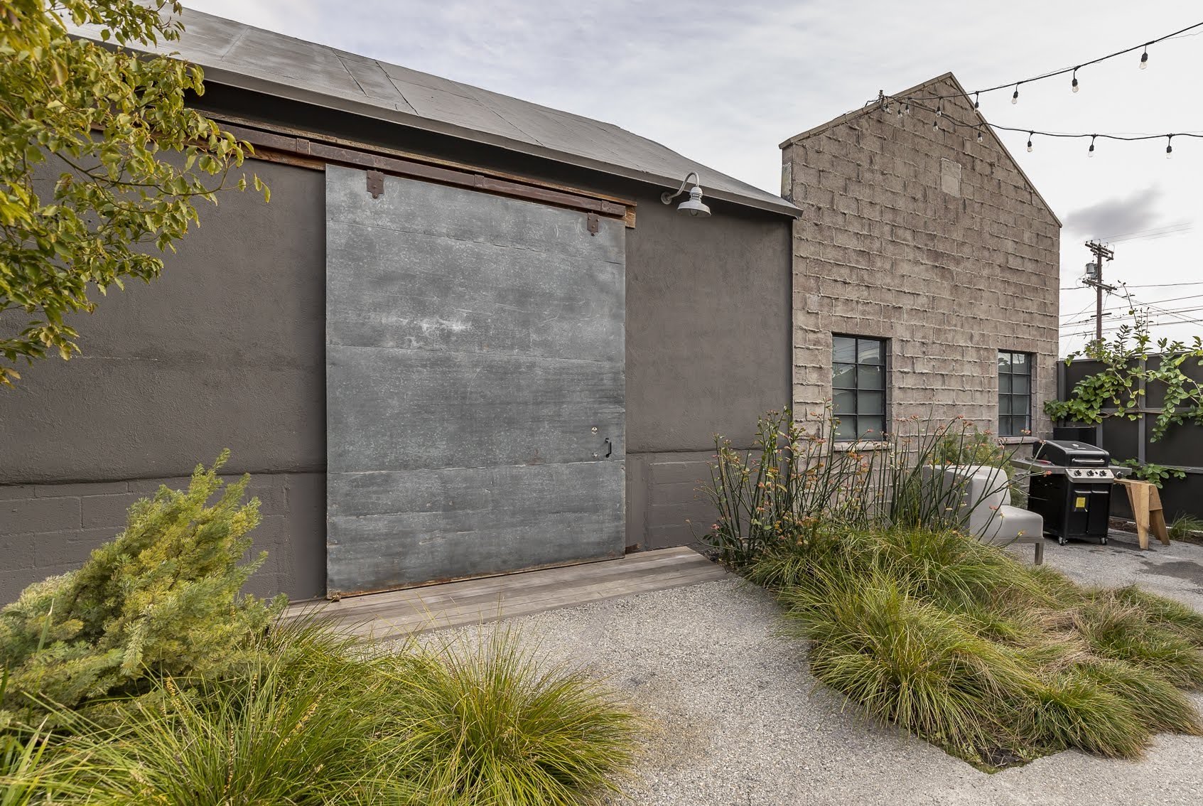 Outdoor patio area with a large gray sliding barn door, brick and concrete walls, plants, a white bench, a barbecue grill, and string lights overhead.