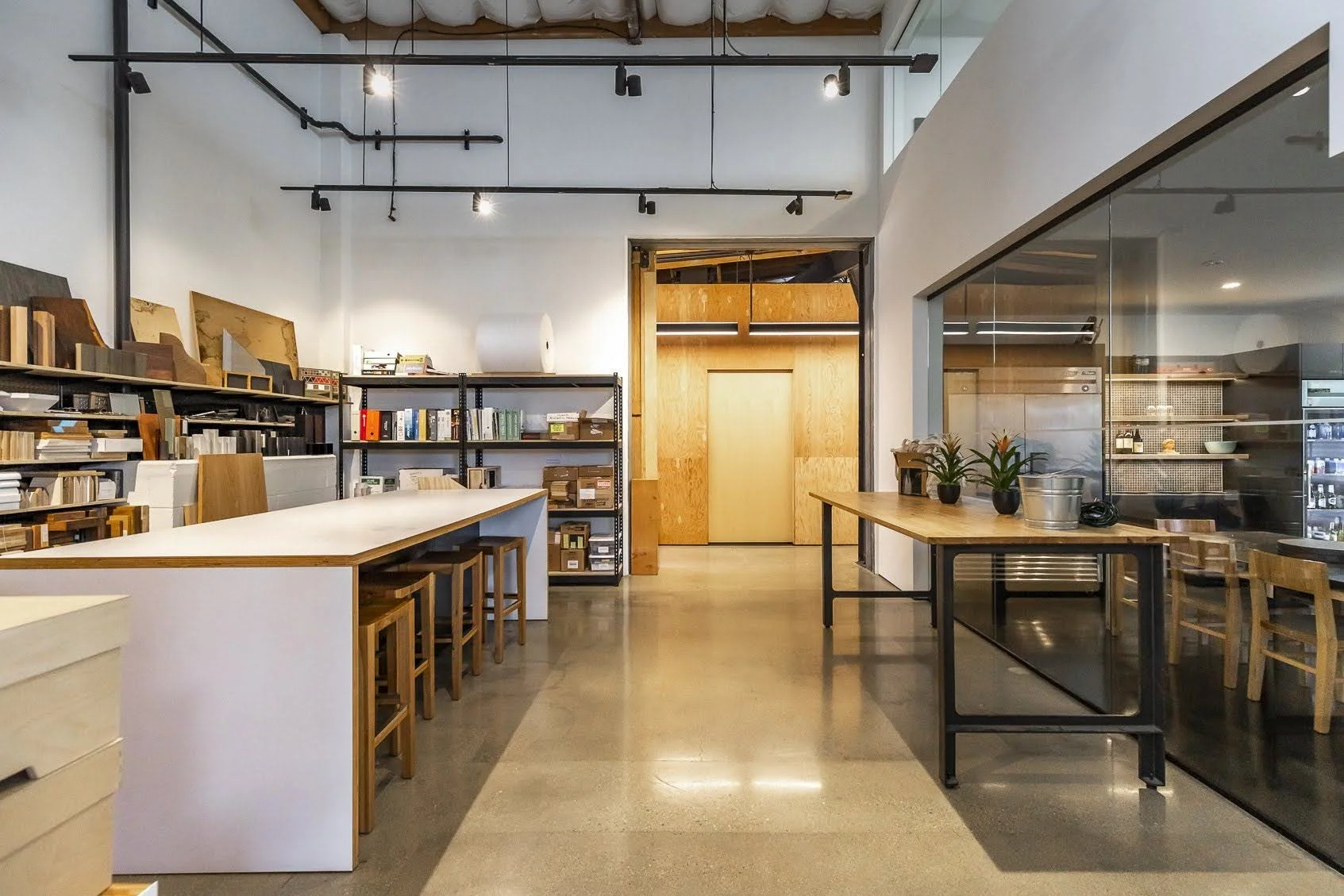 Minimalist interior of a modern workshop or studio space with wooden tables, chairs, bookshelves, and plants, illuminated by track lighting.