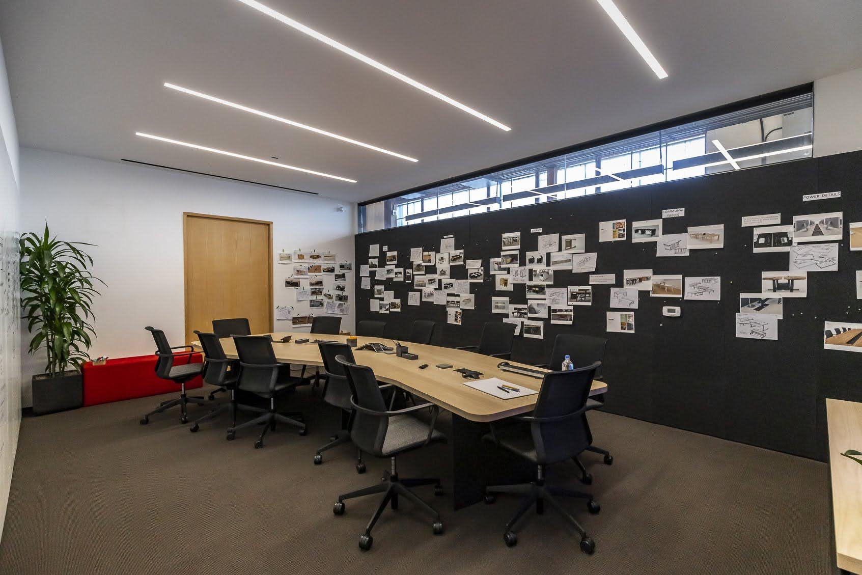 Modern conference room with a large wooden table, black office chairs, a wall decorated with architectural plans and photos, a small water bottle, notebooks, and office supplies, in a well-lit room with ceiling lights and windows.