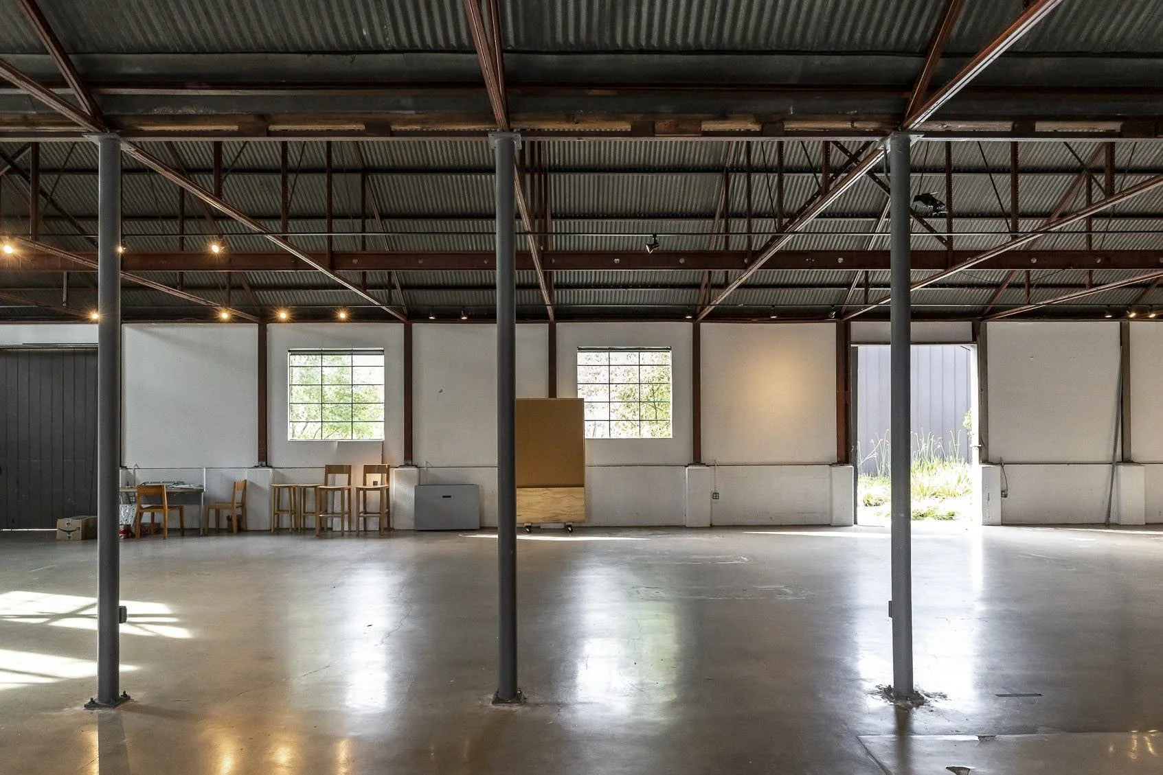 Empty industrial space with metal support beams, concrete floor, and white walls with large windows, some furniture against the wall, and a large open doorway showing outside greenery.
