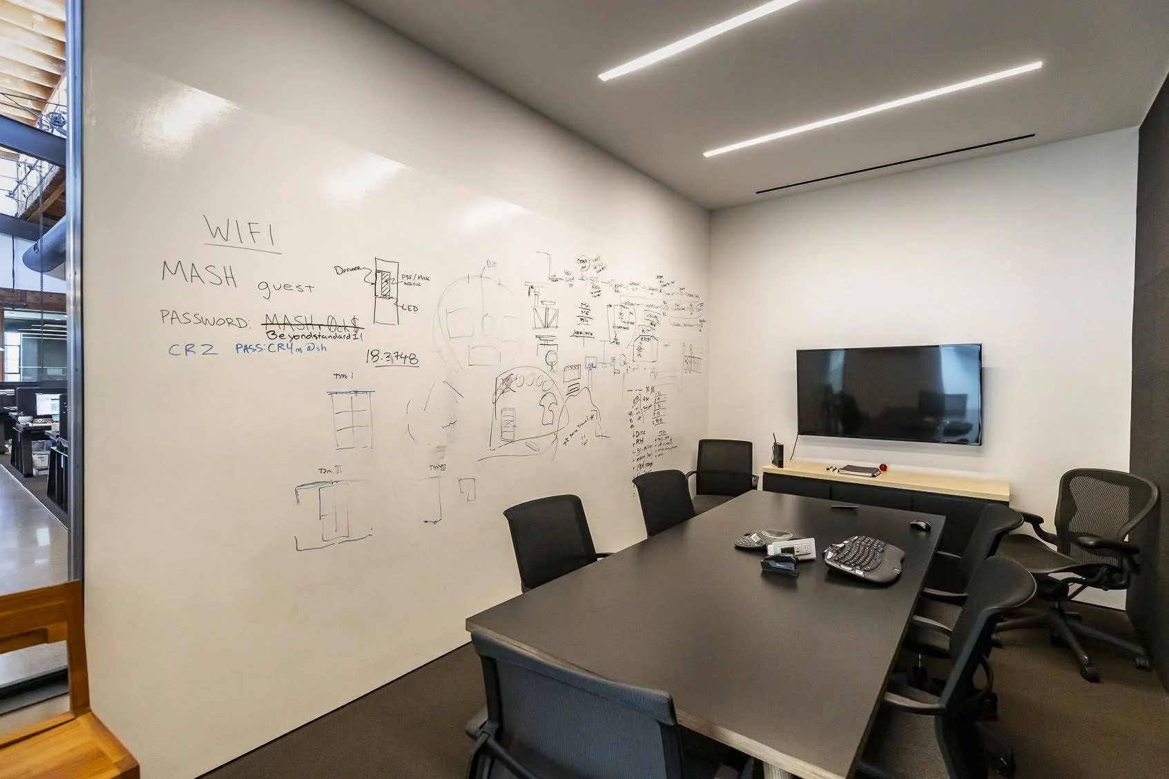 Modern conference room with whiteboard wall filled with sketches and notes, a TV on the wall, and a large table with black office chairs around it.