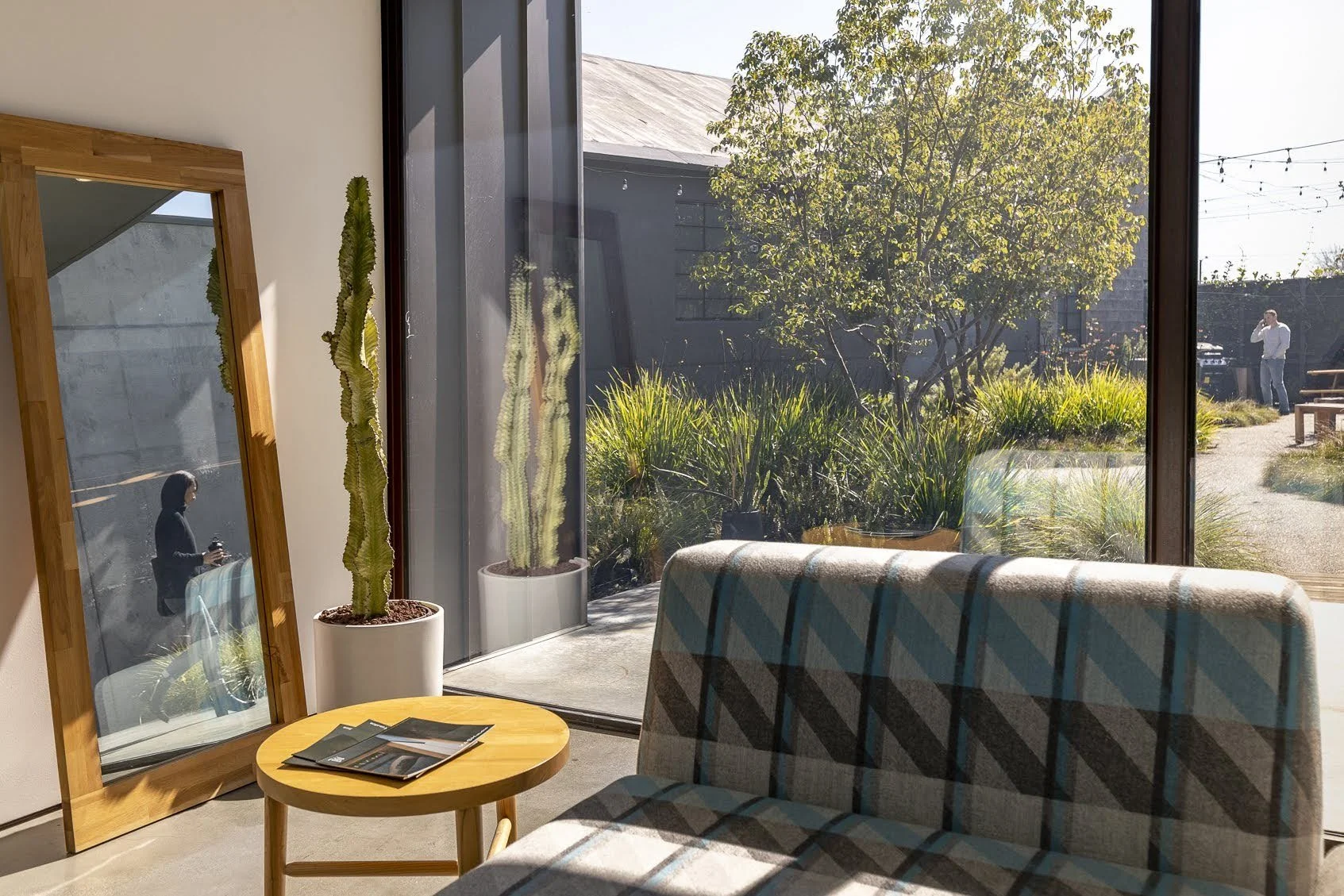 Interior of a room with a large glass window showing a garden outside, potted cactus plants near the window, a round wooden table with magazines, and a sofa with plaid upholstery.