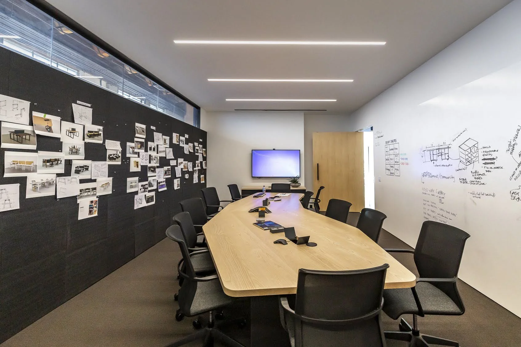 Empty conference room with a large elliptical table, black chairs, a wall with sketches and drawings, a notice board with images, a wall-mounted display, and windows with blinds.