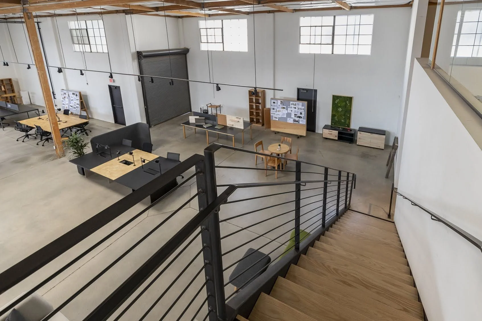 Interior view of a modern office or studio with wooden stairway, large windows, and various tables, workstations, and presentation boards.