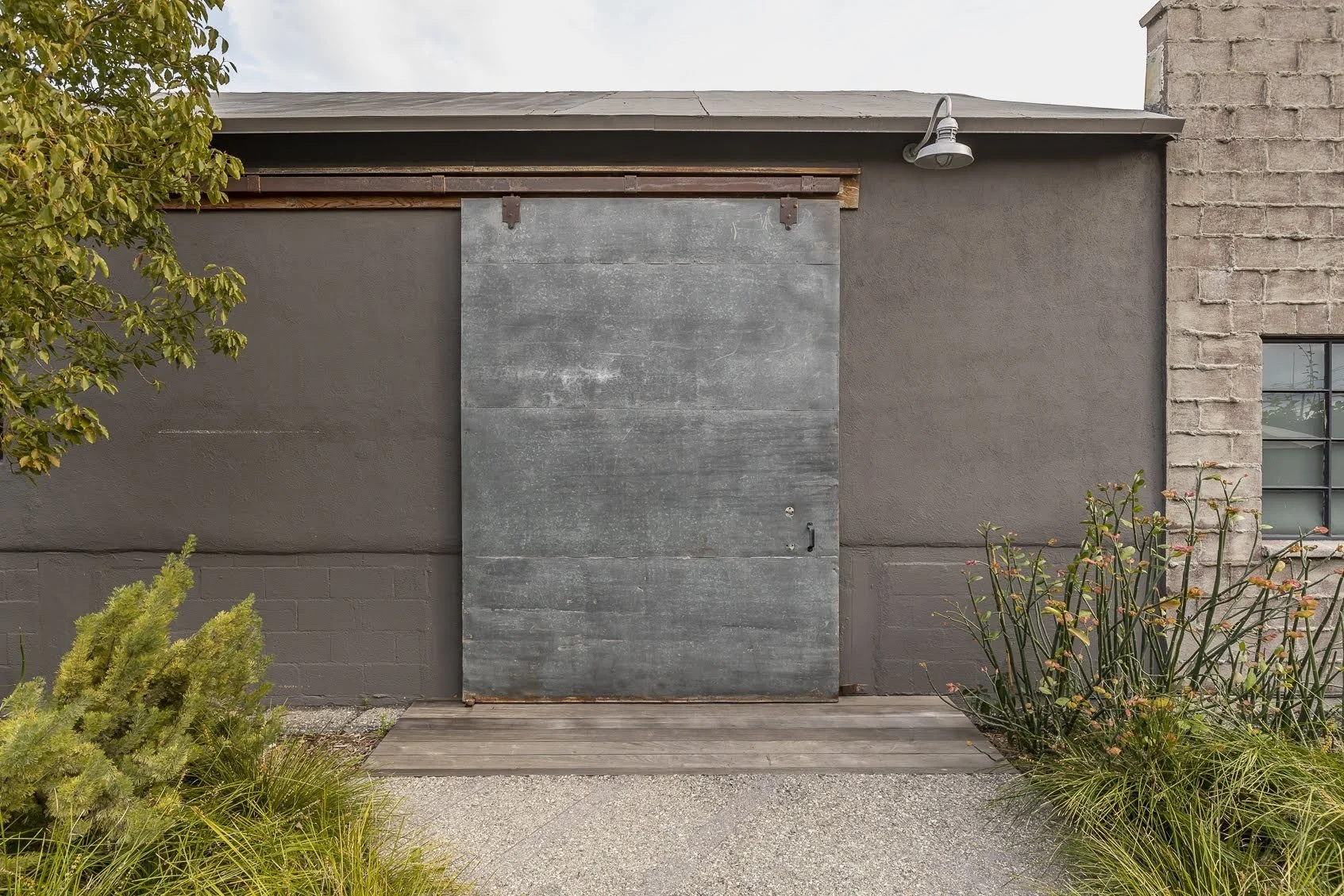 Exterior wall of a building with a large metal sliding door and a small window, surrounded by plants and gravel.