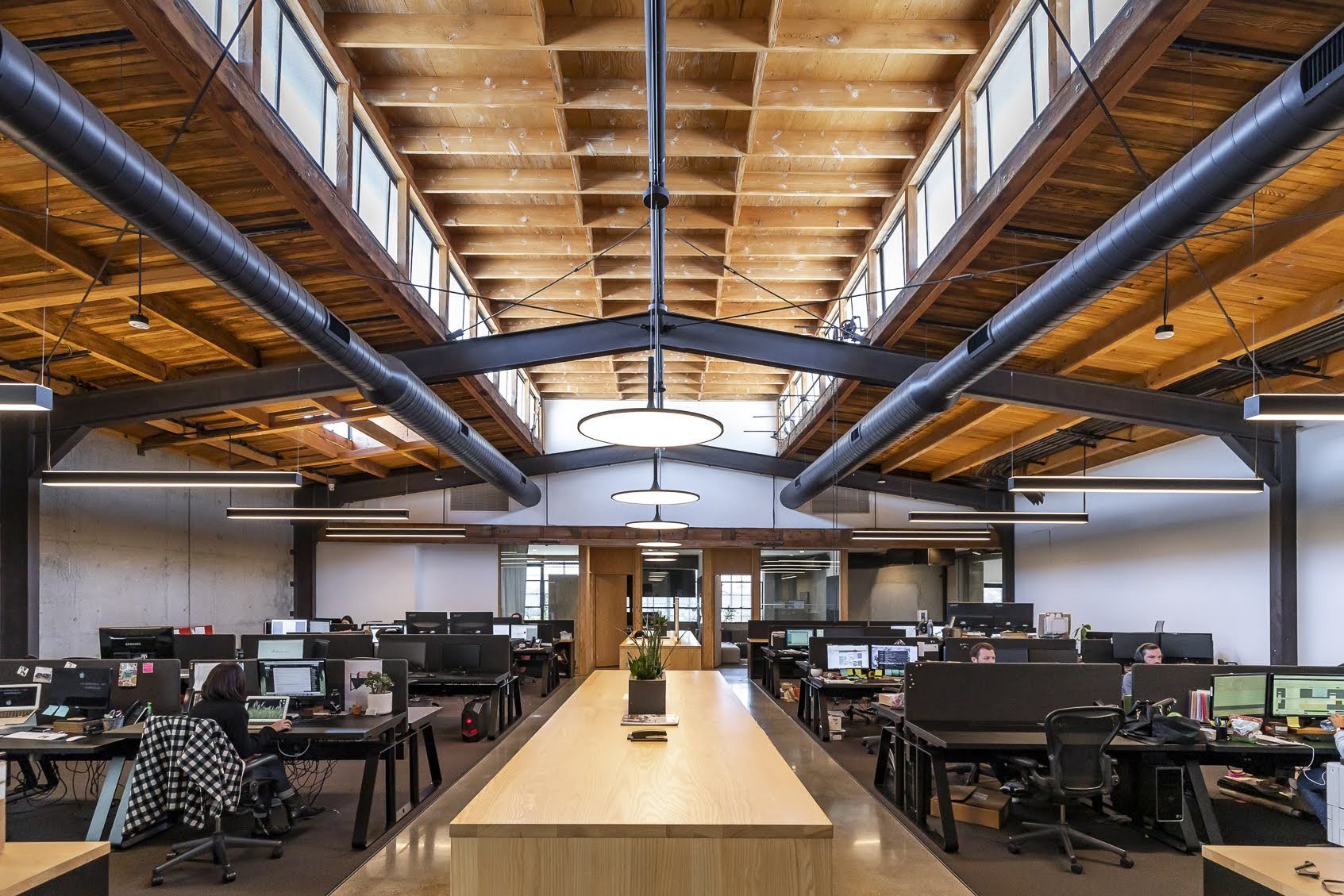 Open-plan office with multiple workstations, black desks, and monitors. Wooden ceiling with large black air ducts and skylights, modern lighting, and a long wooden table with a potted plant in the center.