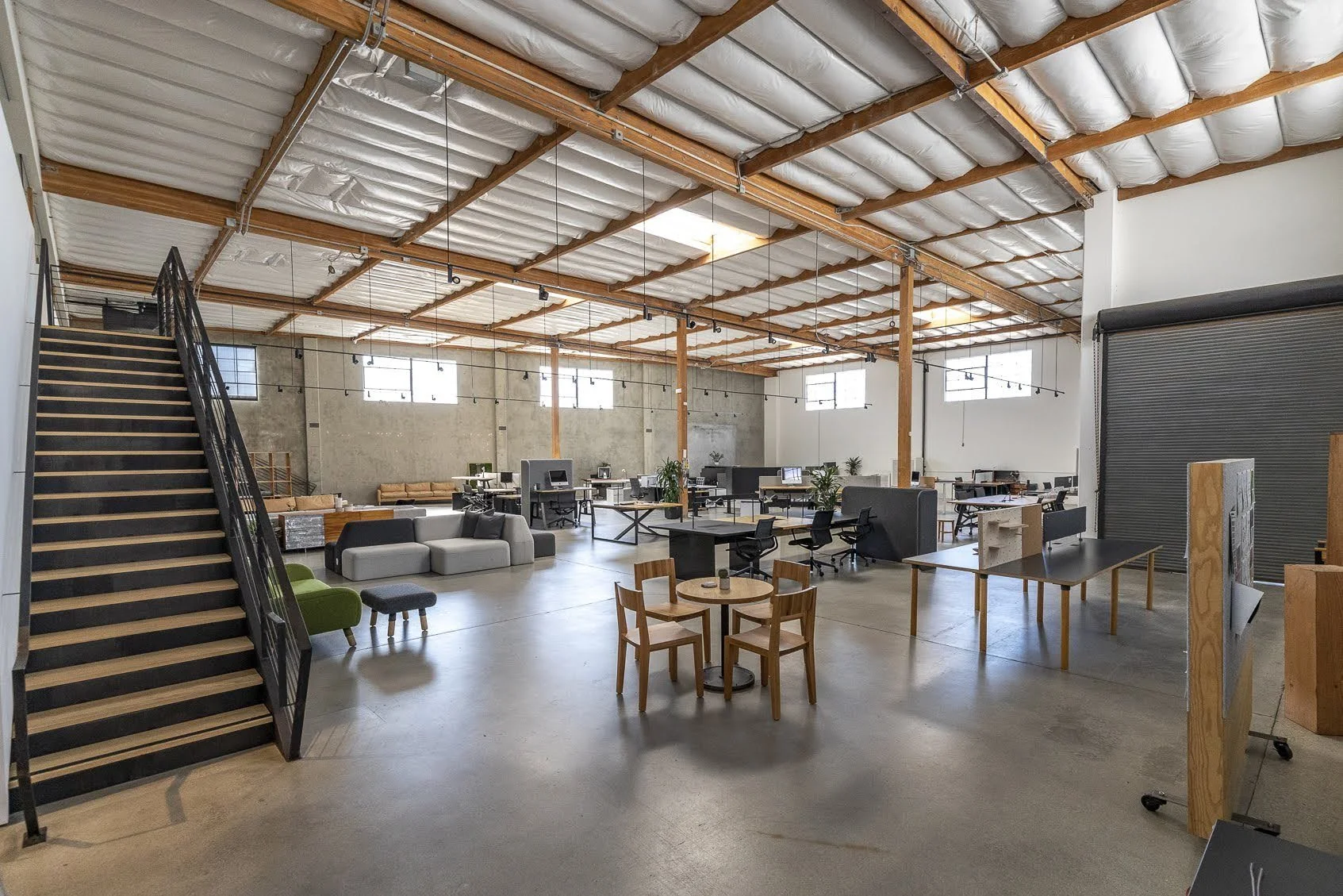 Open industrial-style office space with high ceiling, exposed beams, and large windows. Contains desks with computers, a seating area with a sofa and chairs, and a staircase on the left side. Natural light illuminates the room.