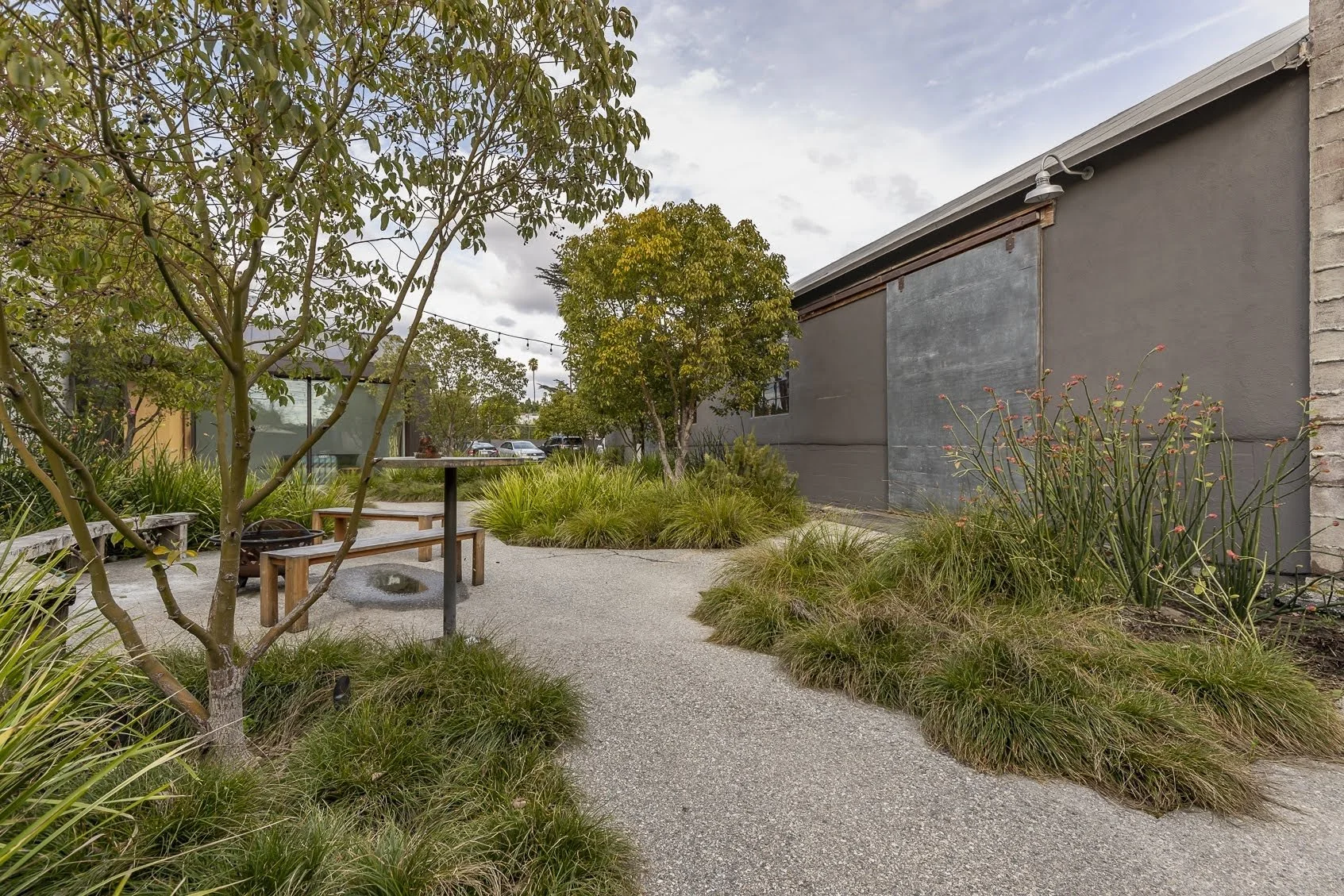 A landscaped outdoor seating area with gravel pathways, trees, ornamental grasses, and benches, next to a gray building under cloudy sky.