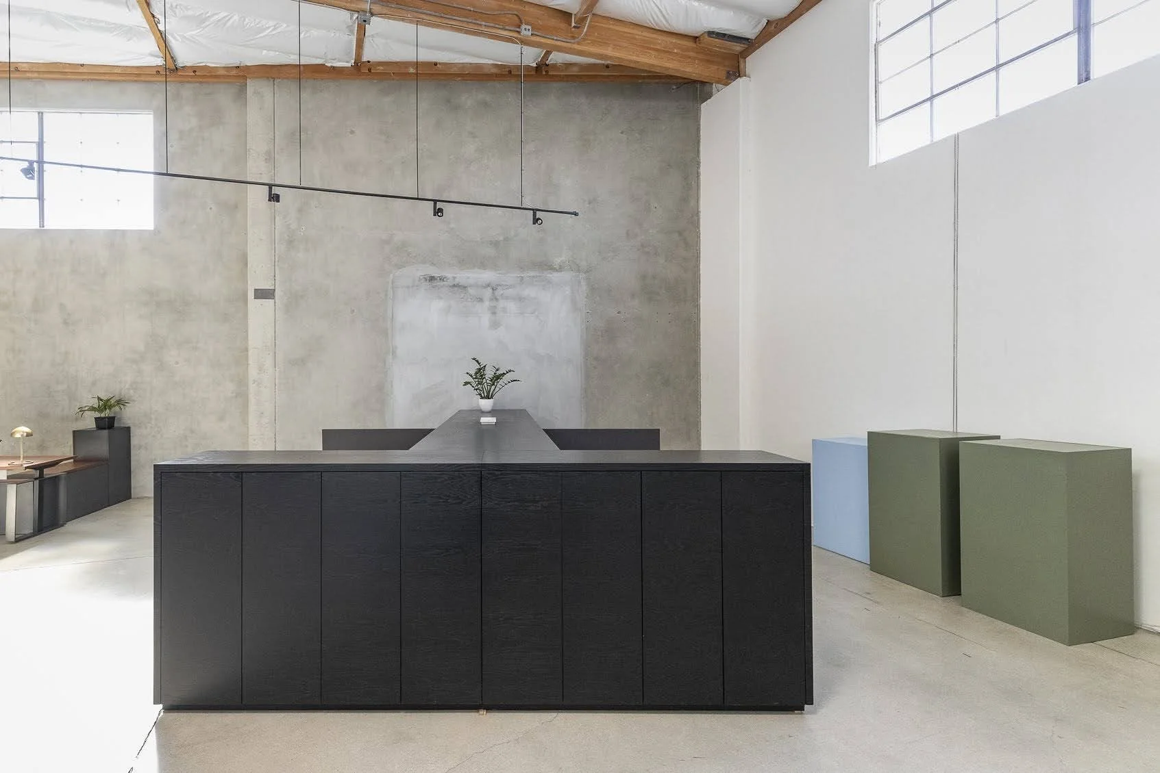 Minimalist interior with a large black cabinet and two cubic pedestals, one light blue and one olive green, in a spacious room with high ceilings, exposed wooden beams, and large windows.