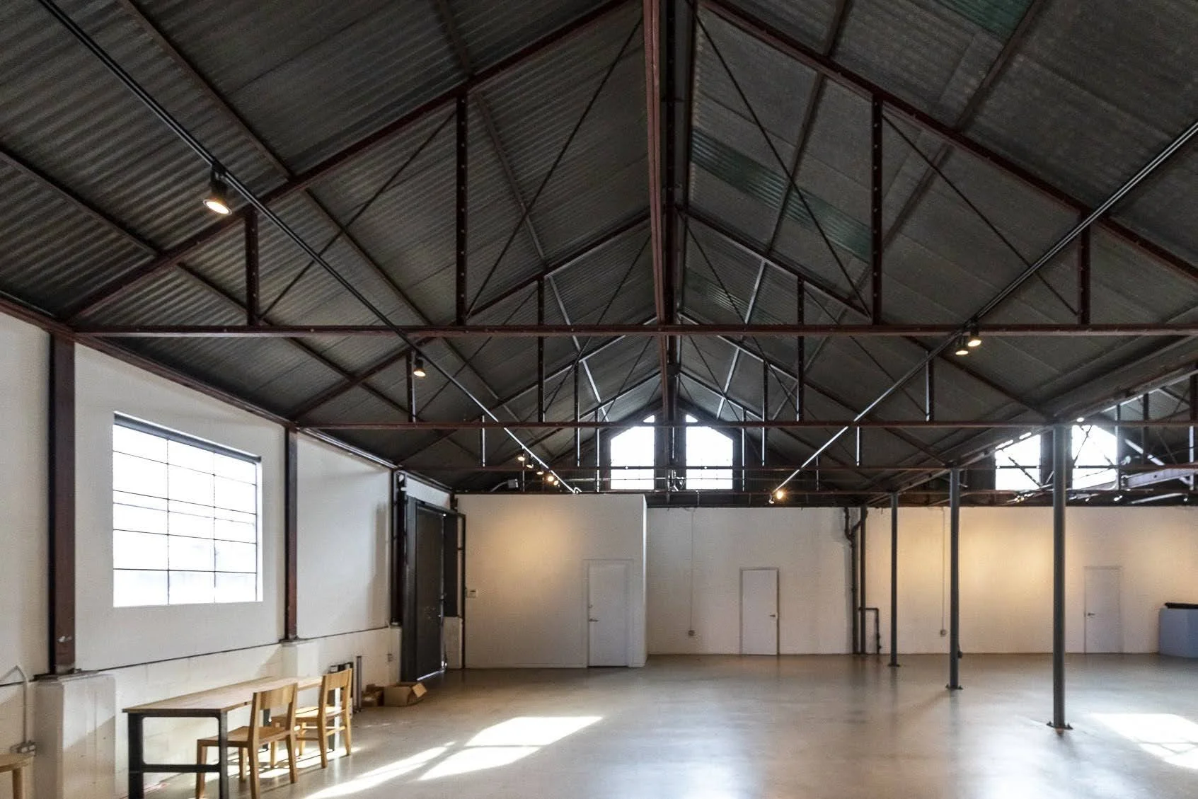 Empty industrial-style room with high metal ceiling, white walls, large windows, and a few chairs along the wall.
