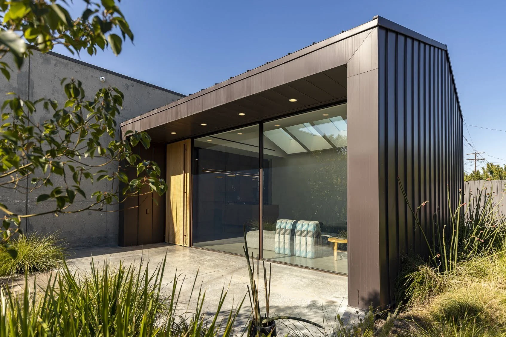 Modern house exterior with glass sliding door, wooden accents, and black metal siding, surrounded by greenery under a clear blue sky.