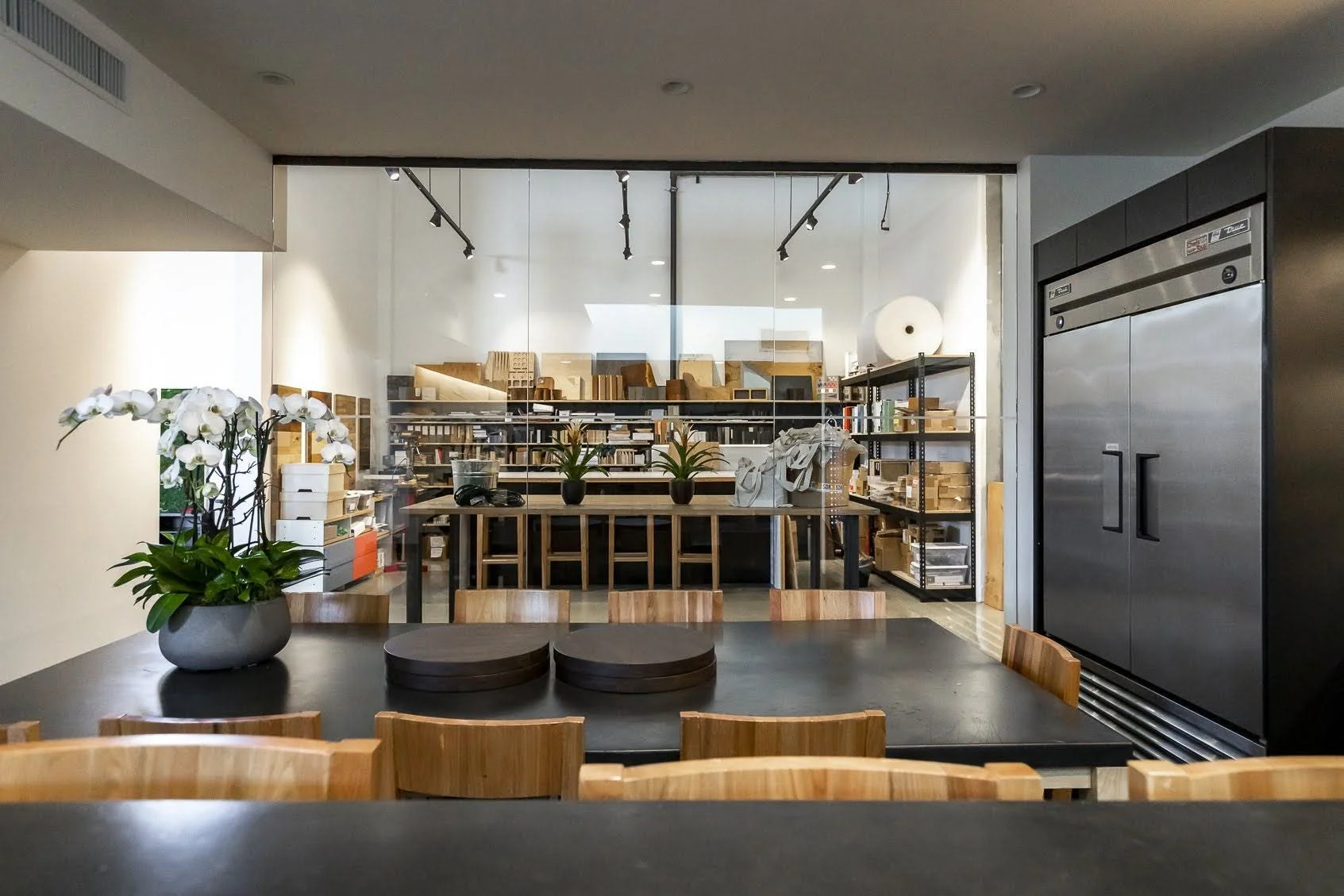 A conference room with a black table, wooden chairs, and a floral centerpiece; behind it, a glass partition reveals a storage room with shelves and boxes.
