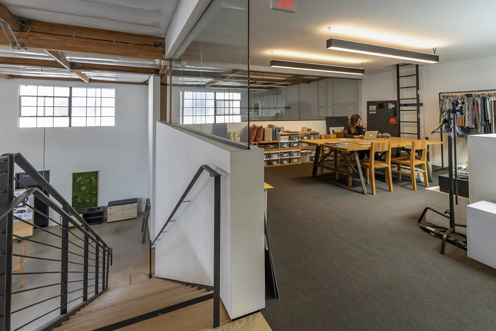 An open-concept office space with a woman working on a laptop at a wooden desk. The room has large windows, a staircase leading down, and various office supplies and decor.