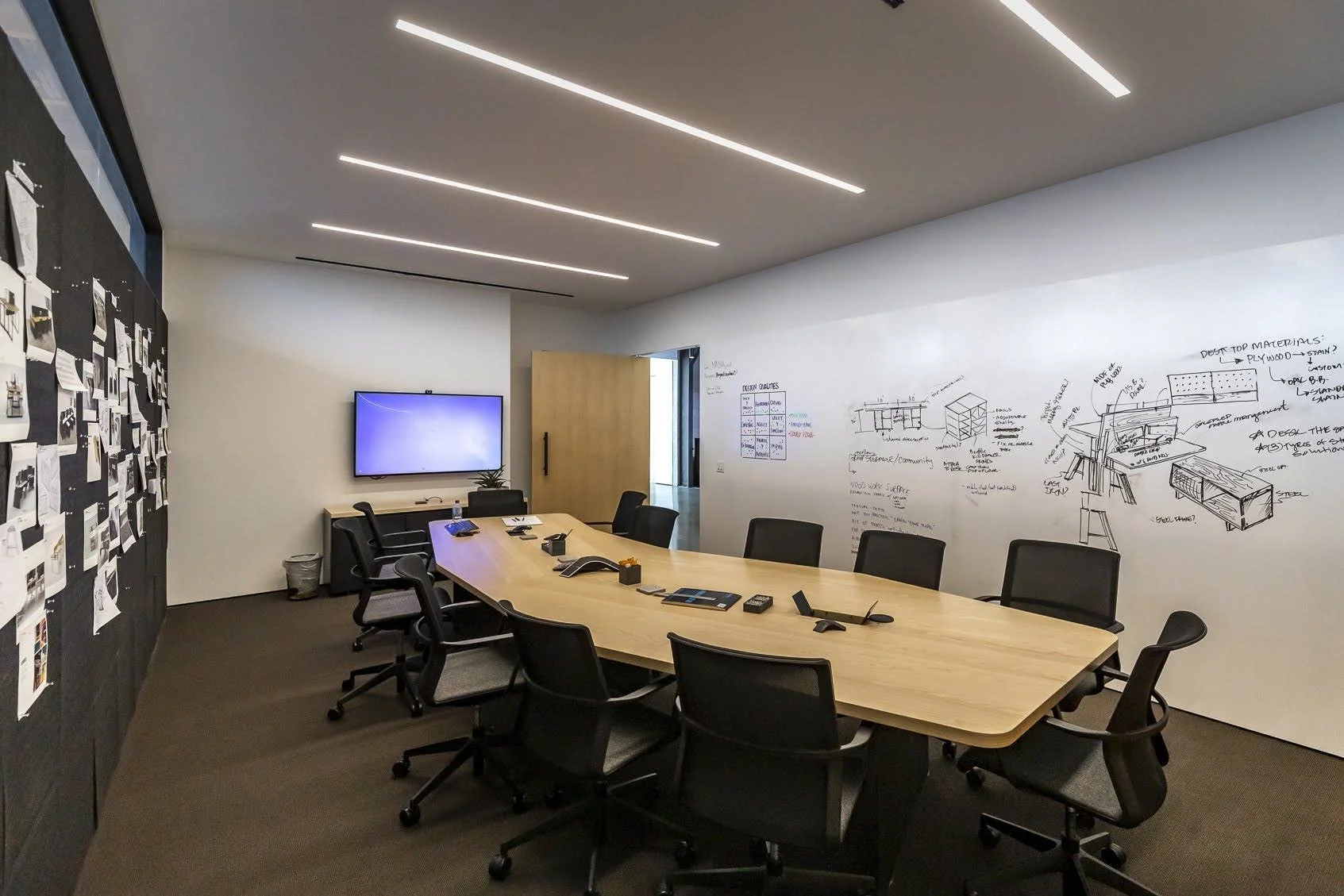Empty conference room with a wooden table surrounded by black office chairs, a large wall with sketches and notes, a whiteboard, and a wall-mounted screen.