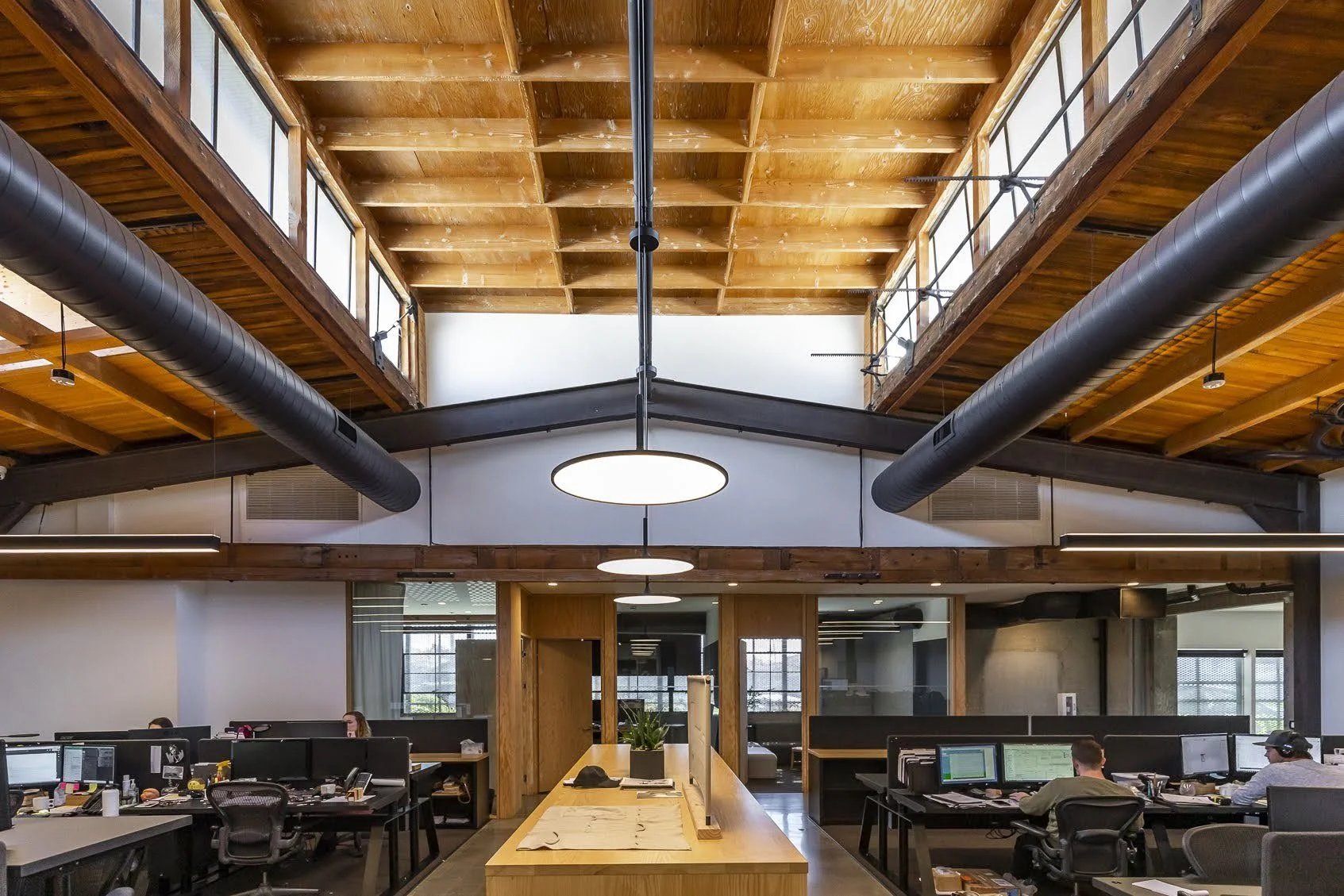 Open-concept office space with wooden ceiling and large black ductwork, featuring multiple computer desks with monitors and employees working.