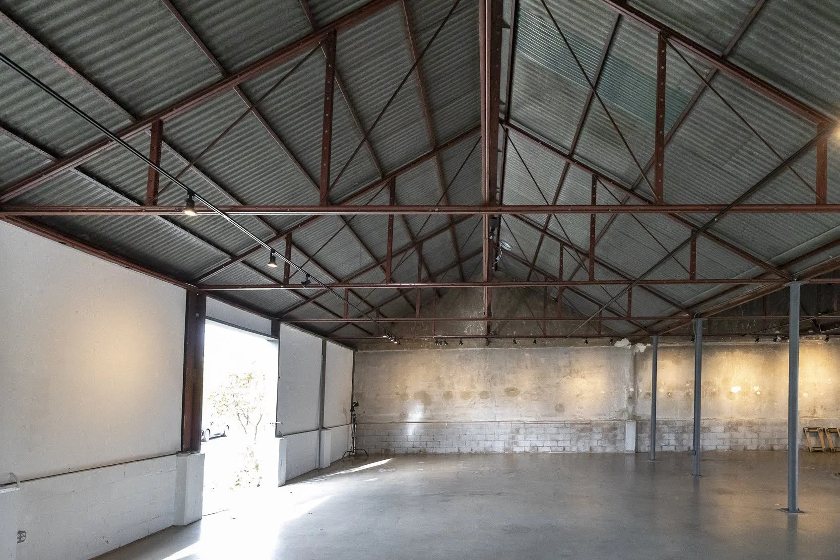 Empty industrial warehouse with metal roof and concrete floor, sunlight entering through open garage door.