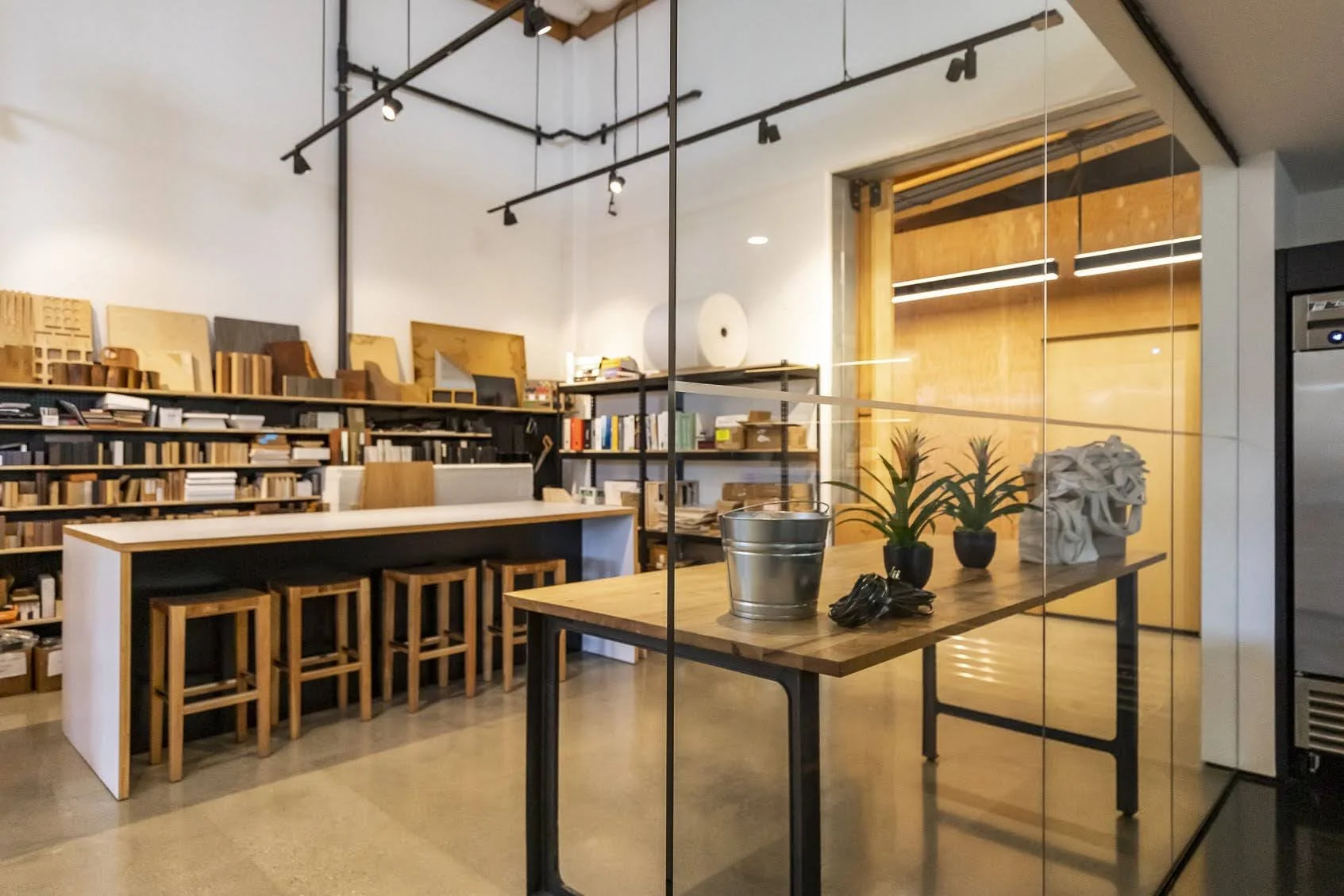 Interior of a modern workshop or studio with a large worktable, wooden shelves filled with books and samples, and outdoor light coming through large windows.