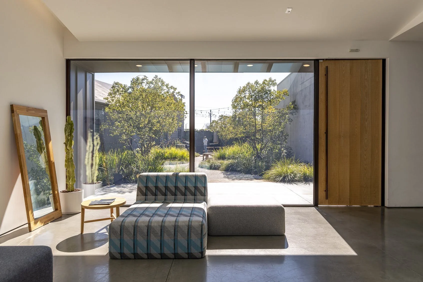 Modern minimalist living room with large glass sliding door overlooking a garden with trees, bushes, and pathway. Contains a plaid upholstered chair, a gray ottoman, a small wooden side table with magazines, and a mirror leaning against the wall with